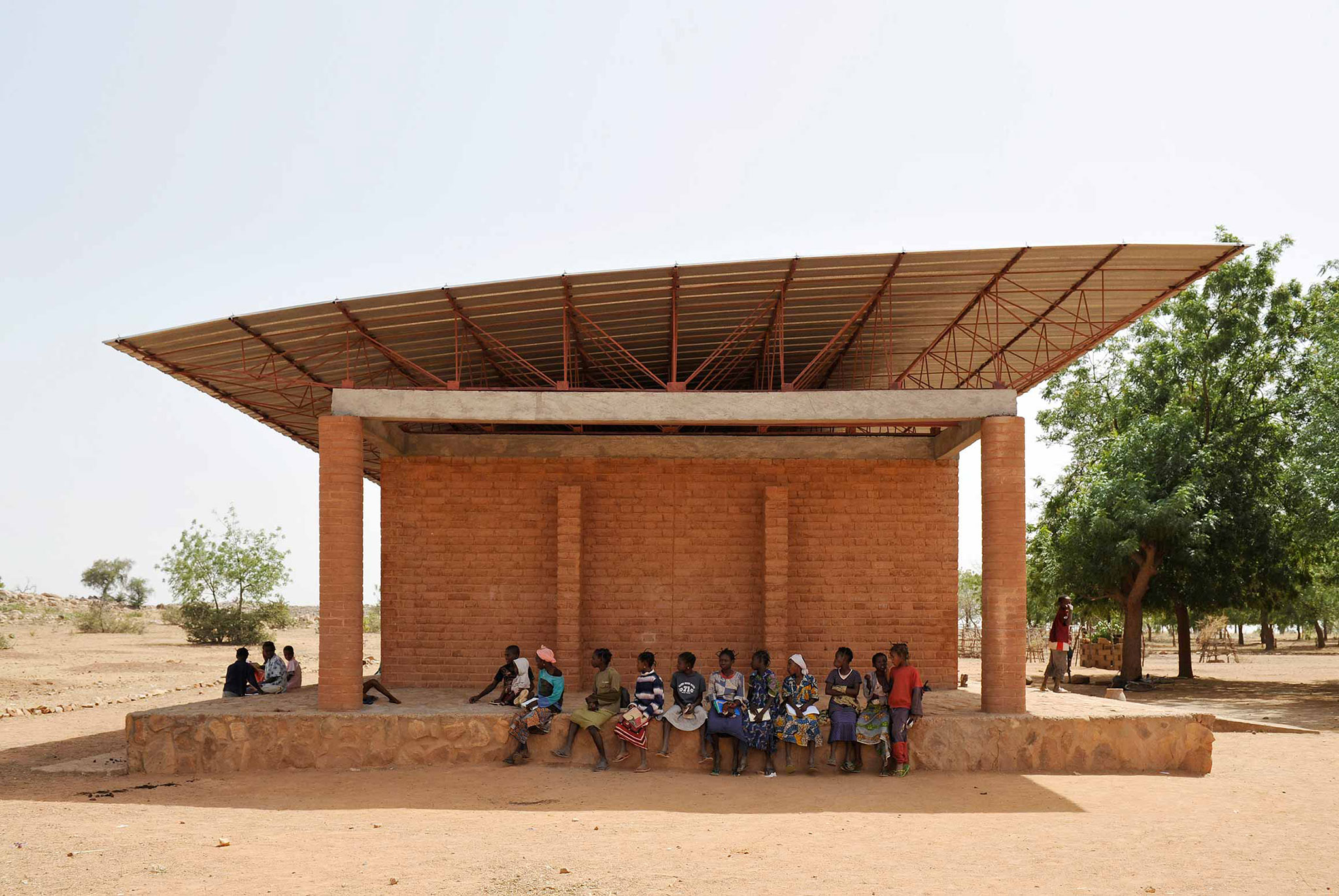 A group of people sit in the shade of a small brick building with a metal roof in a dry, rural area with sparse trees, evoking scenes discussed in MOS’s New Book On Public Spaces.