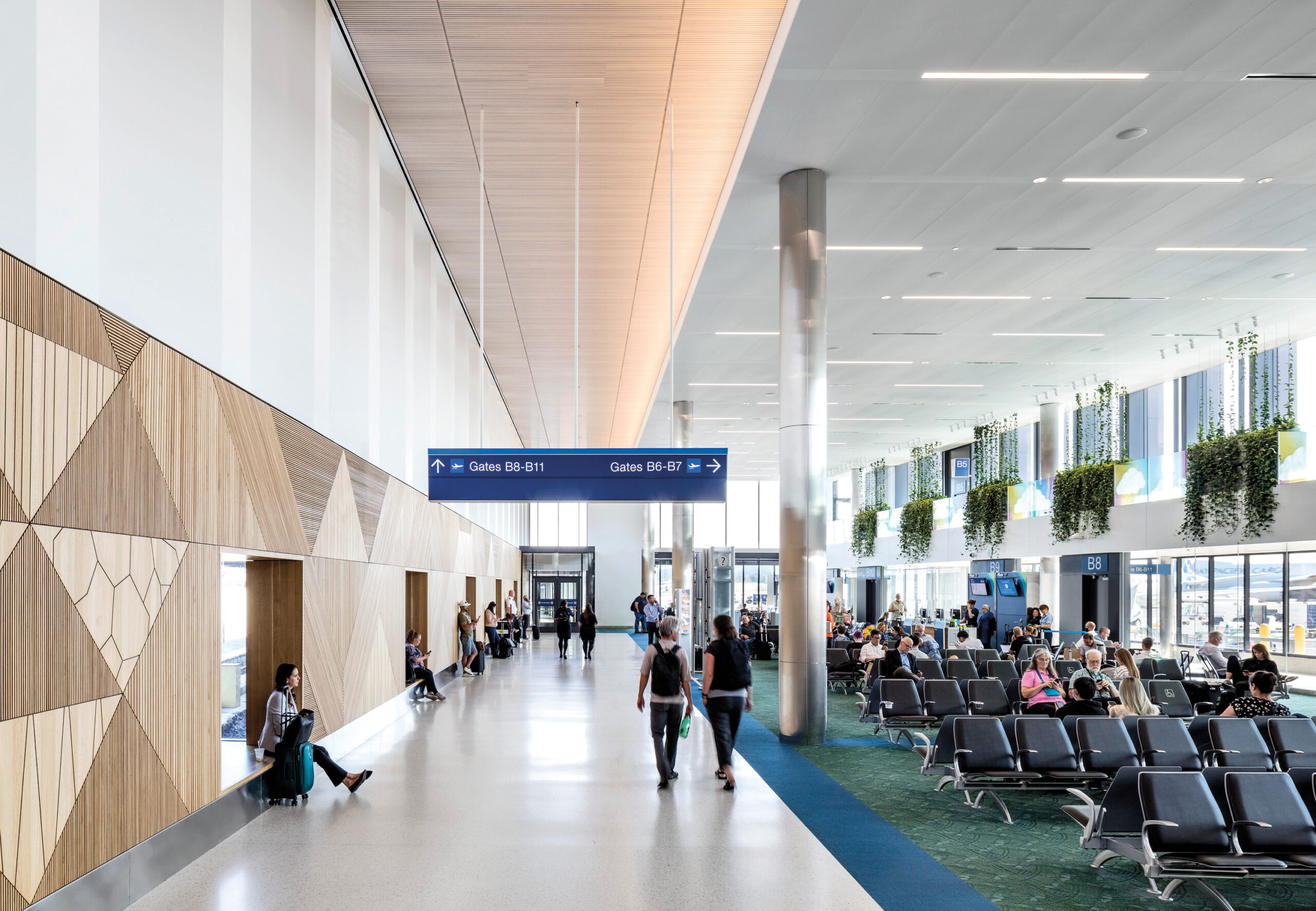 Passengers sit and walk in a modern airport terminal with geometric wall panels, tall ceilings, large windows, and gate signs overhead.