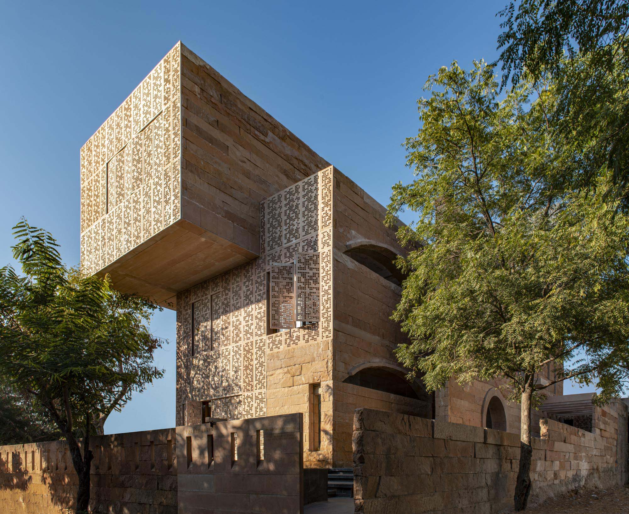 A modern building with patterned stone facades, large cantilevered section, and arched openings, surrounded by trees under a clear blue sky, showcases innovative use of natural stone materials.