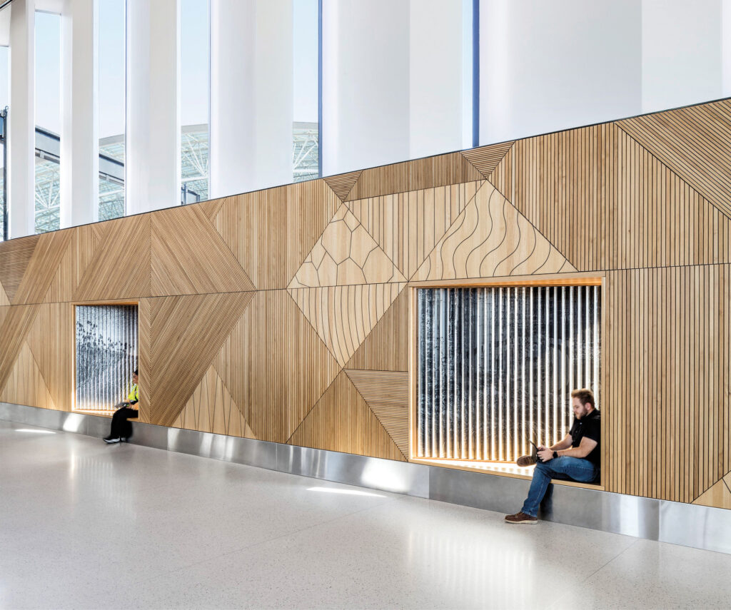 Two people sit in recessed window alcoves of a modern building with geometric wooden wall panels and tall vertical windows.