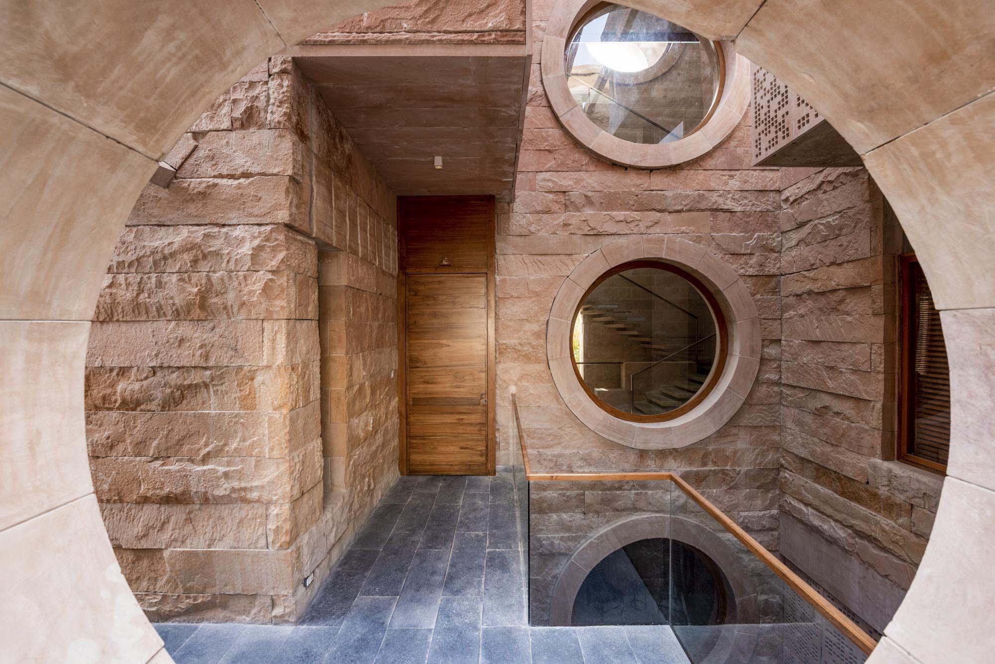 Interior space with stone walls, circular windows, a wooden door, and a dark stone floor featuring a round opening that reflects the surrounding stone architecture above.