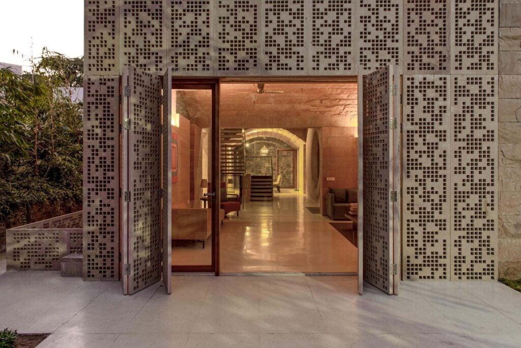 Entrance with open glass doors framed by perforated stone panels, revealing a modern interior with elegant stone arches, comfortable seating, and warm lighting.
