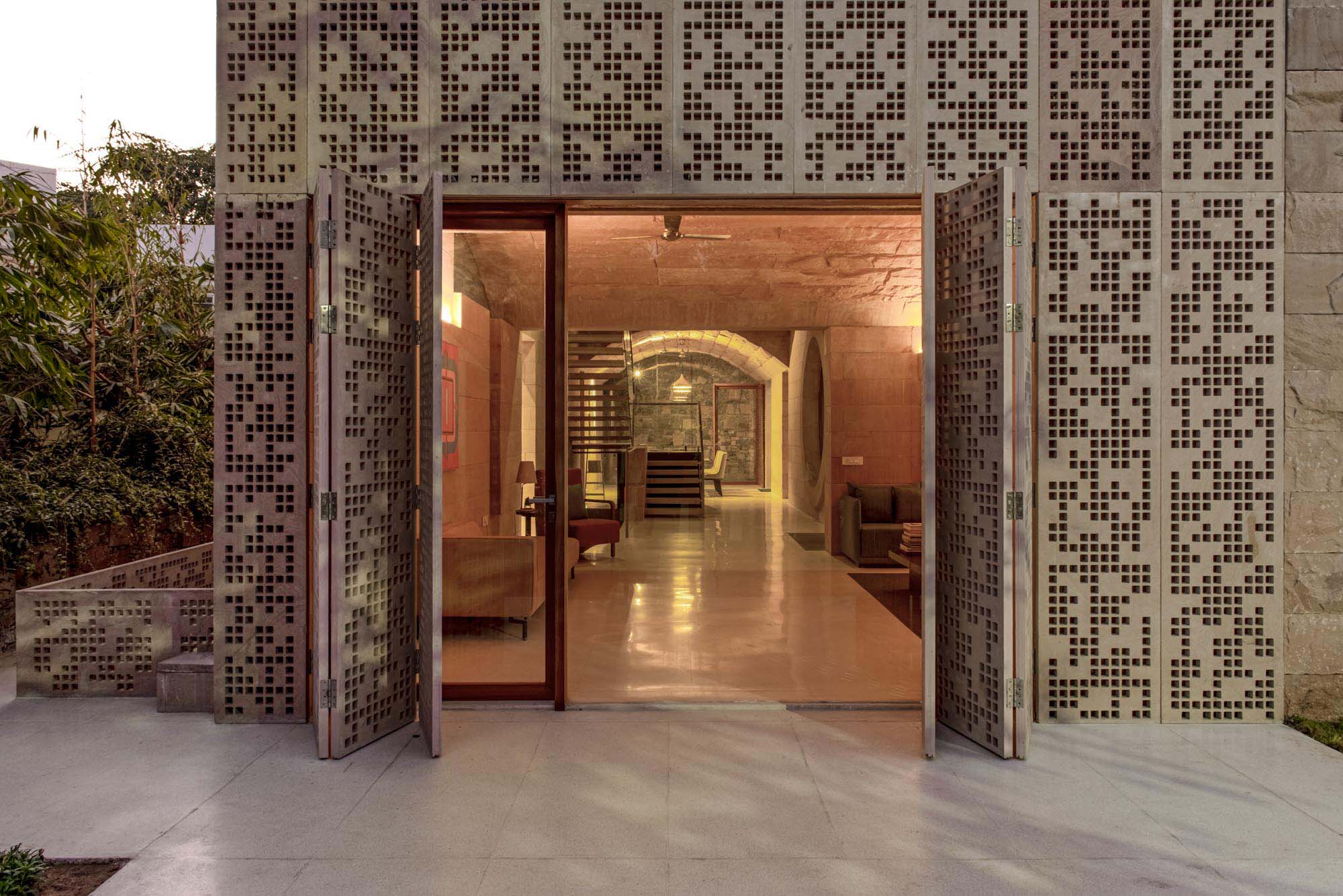 Entrance with open glass doors framed by perforated stone panels, revealing a modern interior with elegant stone arches, comfortable seating, and warm lighting.