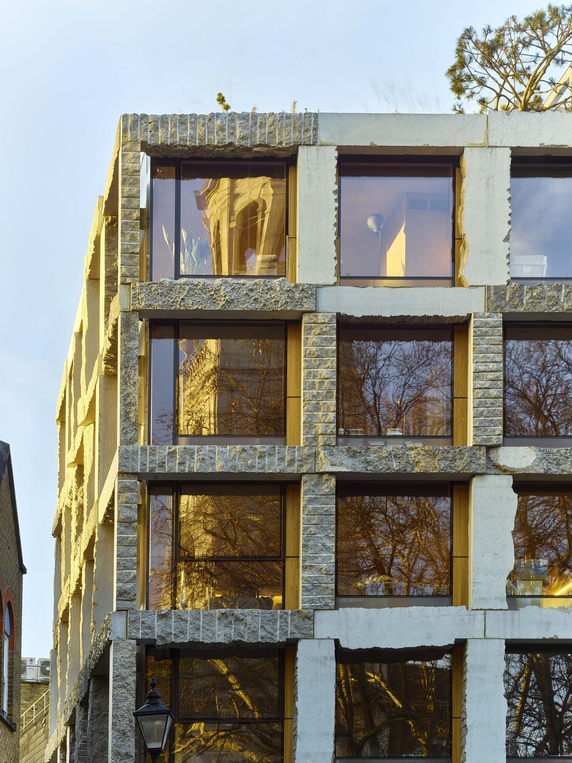 A modern building with large windows framed by rough stone and concrete, reflecting trees and a distant tower in the shimmering glass.