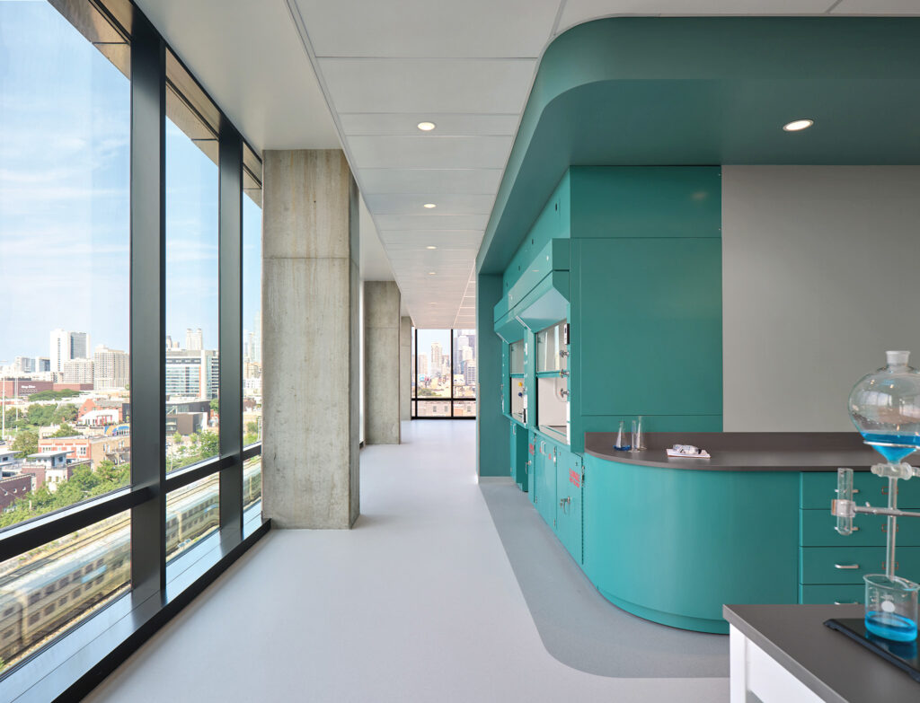 Modern laboratory interior with turquoise cabinetry, large windows overlooking a cityscape, and scientific glassware on the counter.