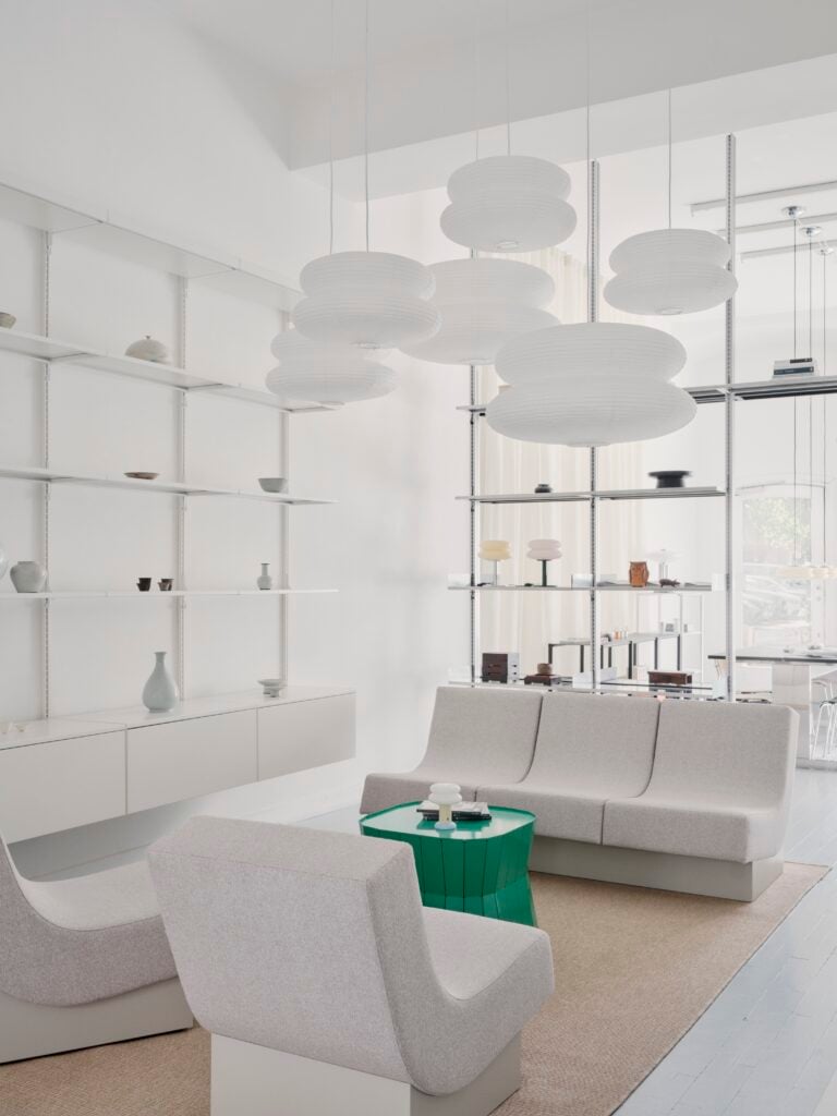 Minimalist living room with light gray sofas, a green geometric coffee table, and wall-mounted shelving is inspired by the latest trends from Milan Design Week 2026, featuring multiple round white pendant lights hanging from the ceiling.