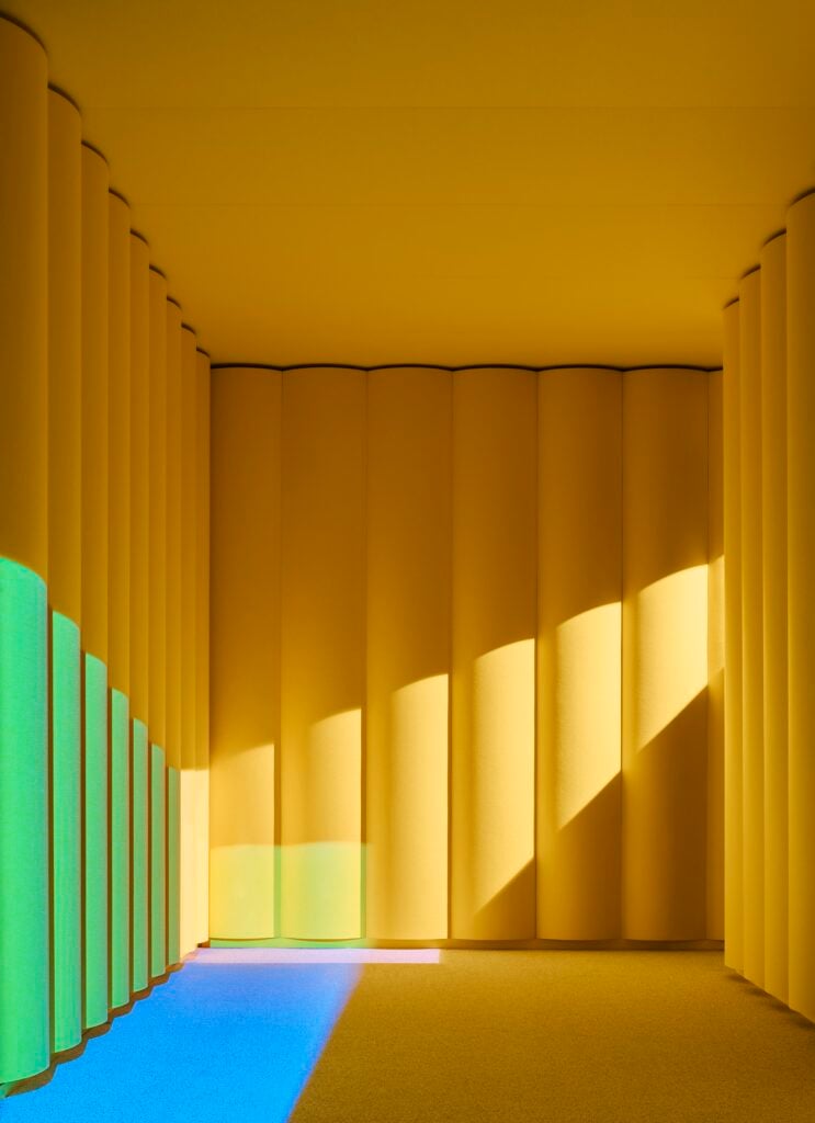 A yellow room with tall cylindrical columns, illuminated by sunlight; colored light from office products casts green and blue shapes on the floor and wall.