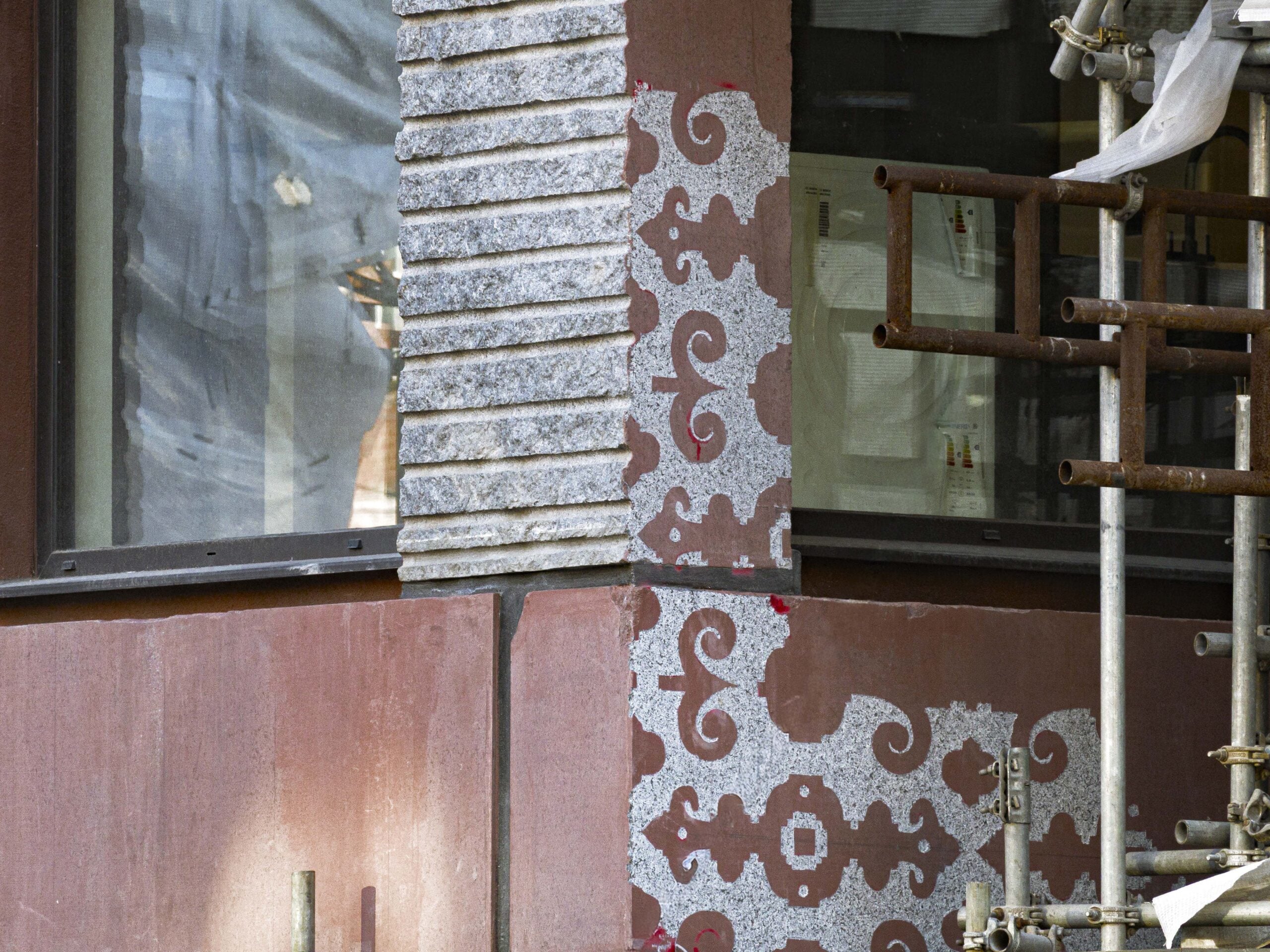 Close-up of a building corner under renovation, showing exposed patterned stonework, ribbed stone paneling, red wall, scaffolding, and reflections in the glass window.