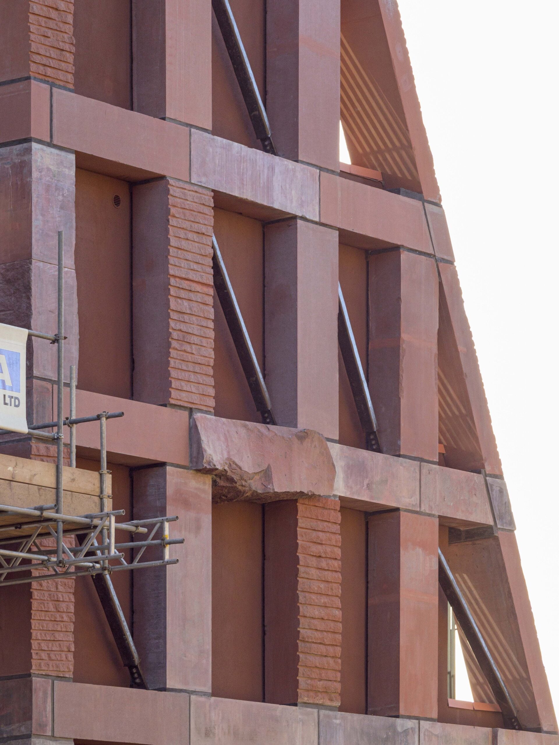 Close-up of a building under construction with exposed steel beams, red brick and concrete sections, and scaffolding on the left side, alongside partially set stone detailing.