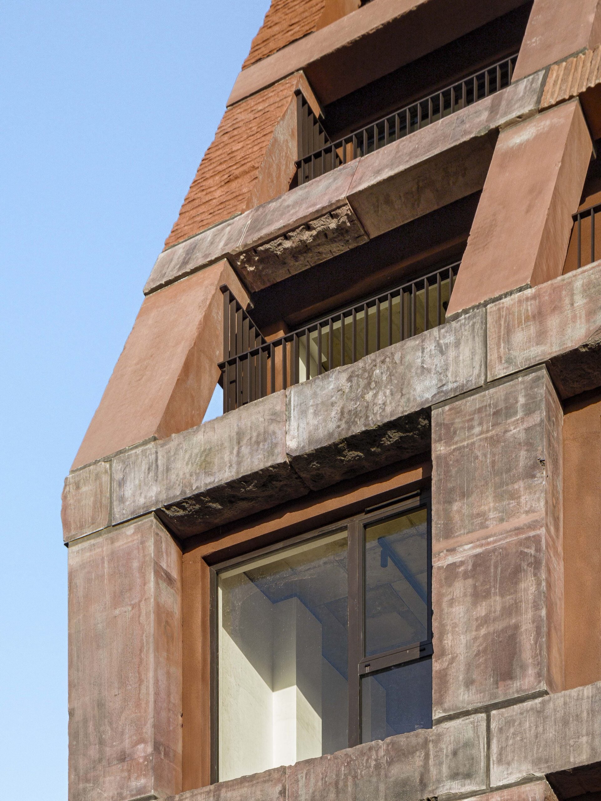 A close-up view of a modern building facade with angular, geometric shapes and a large window, featuring reddish-brown concrete, stone accents, and sleek metal railings.