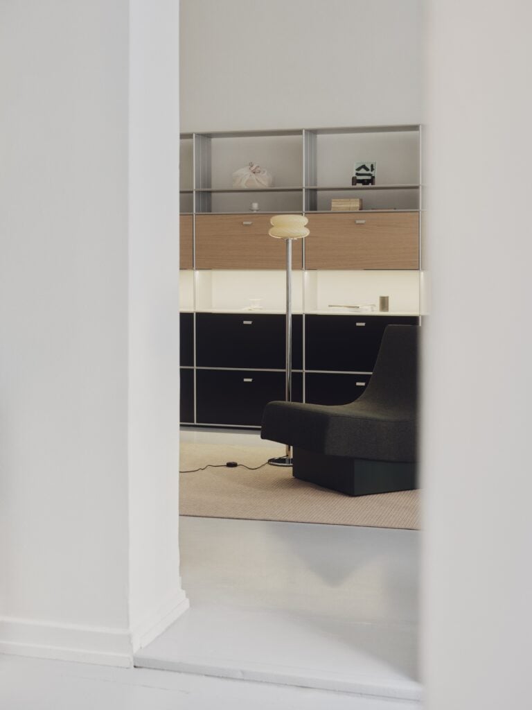 A modern minimalist living room, inspired by Milan Design Week 2026, features a black chair, floor lamp, cabinet shelves, and a beige rug, all artfully viewed through a doorway.