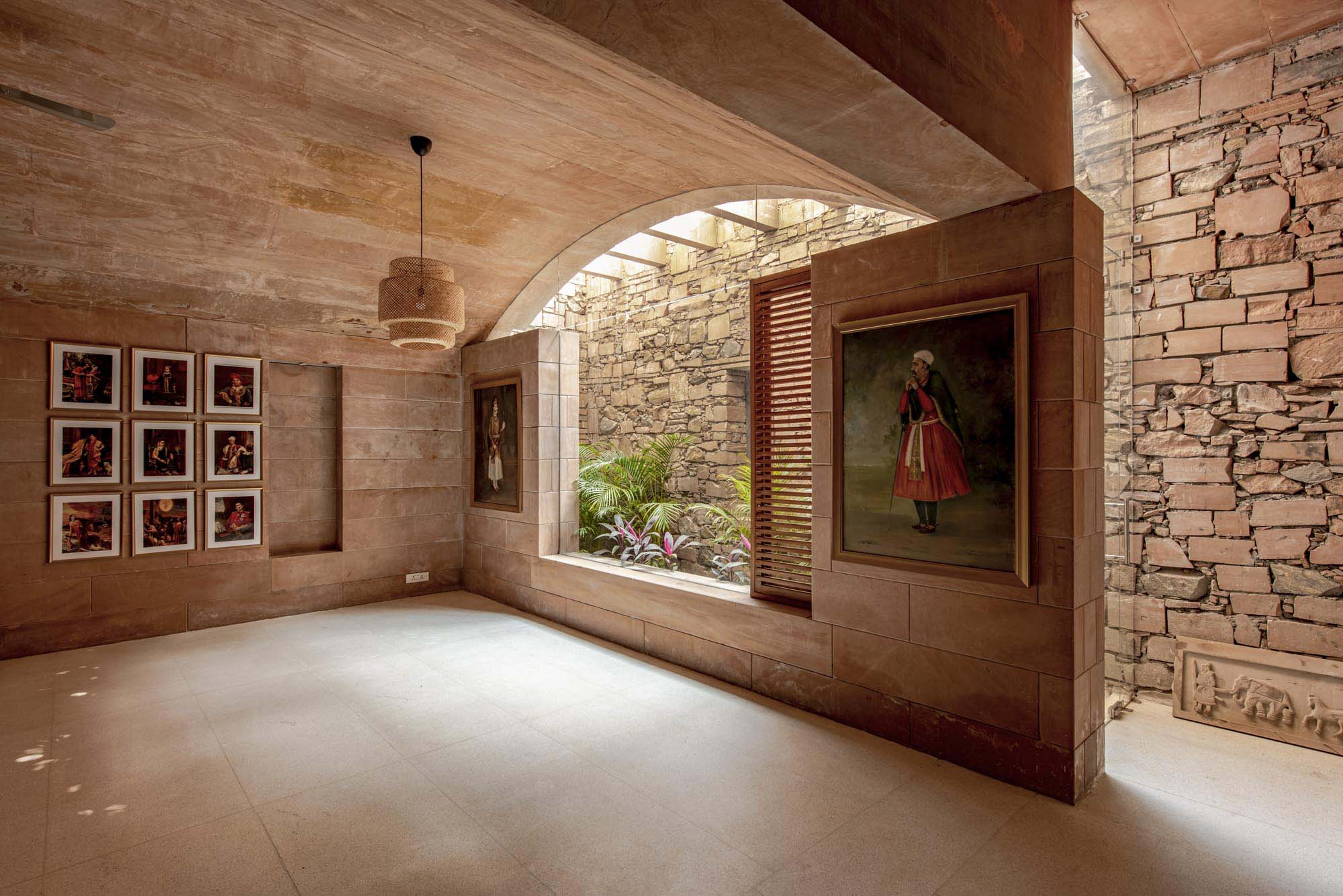 A stone-walled room with framed art, an arched window opening to a small garden, sunlight streaming through slatted shutters, and a rustic stone floor beneath a hanging light fixture.