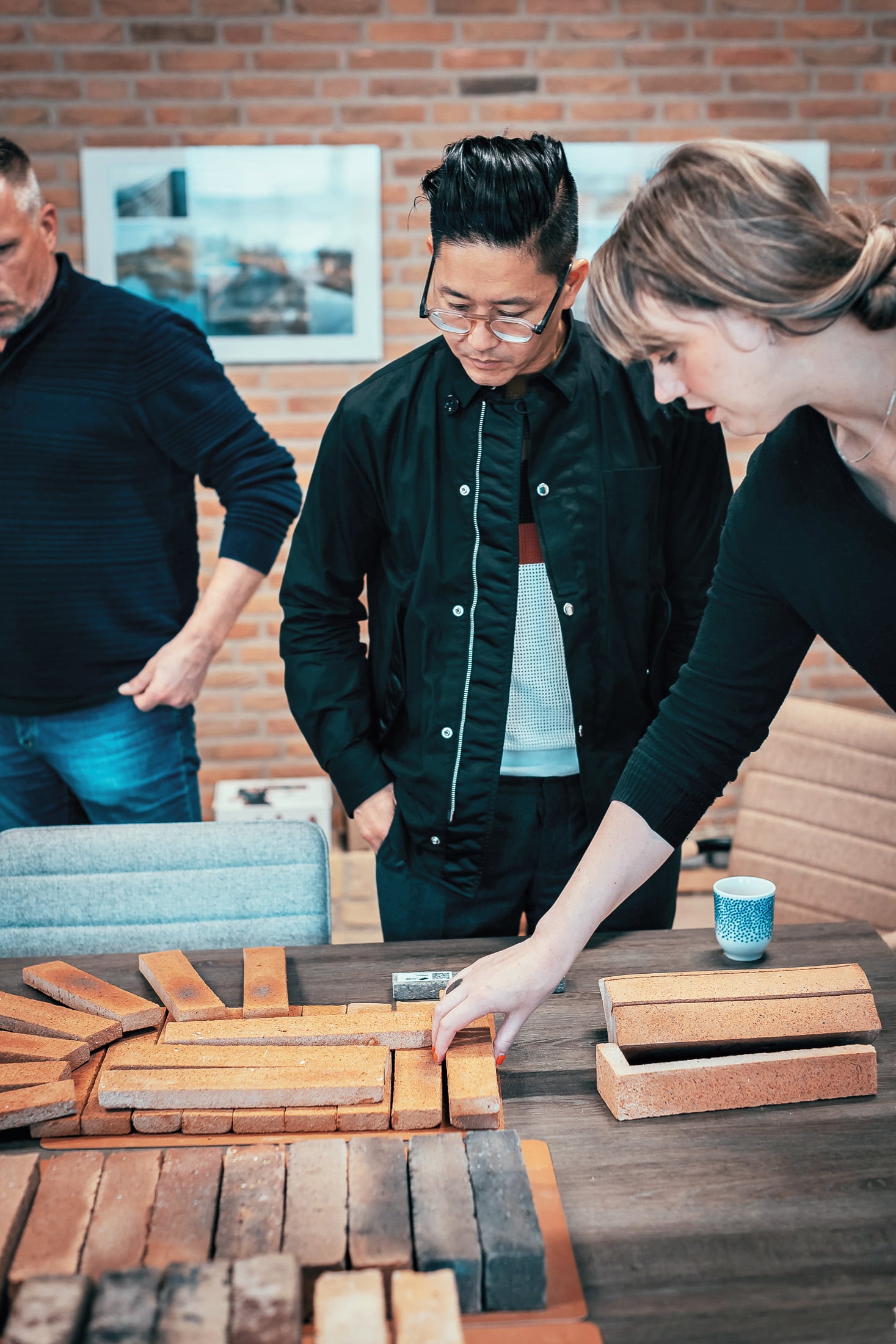 Three people examine and arrange various rectangular bricks on a table in a room with a brick wall and framed photos in the background.