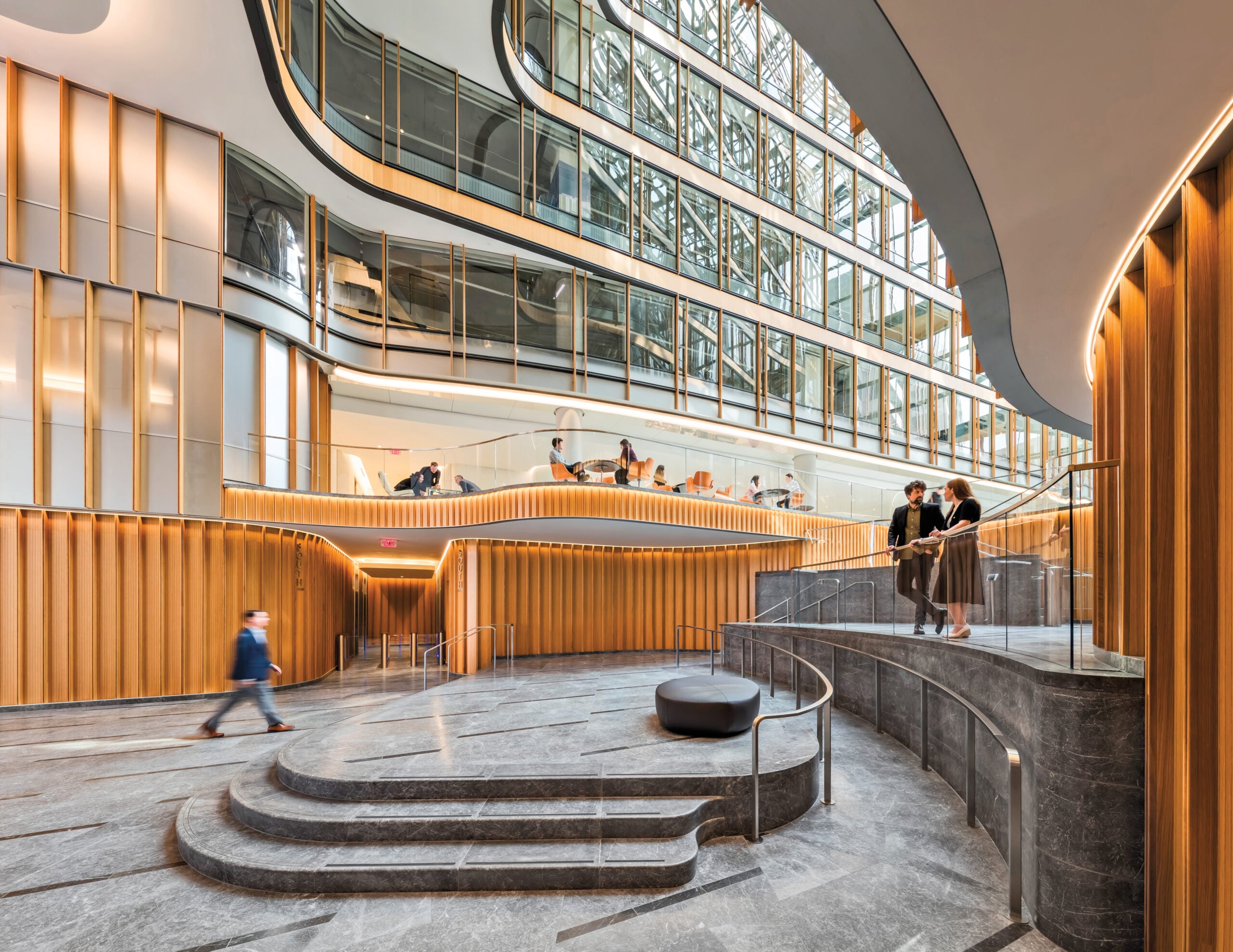 Modern building interior with curved architecture, wood paneling, large windows, and people walking or talking in the spacious, multi-level atrium showcasing craft in architecture.