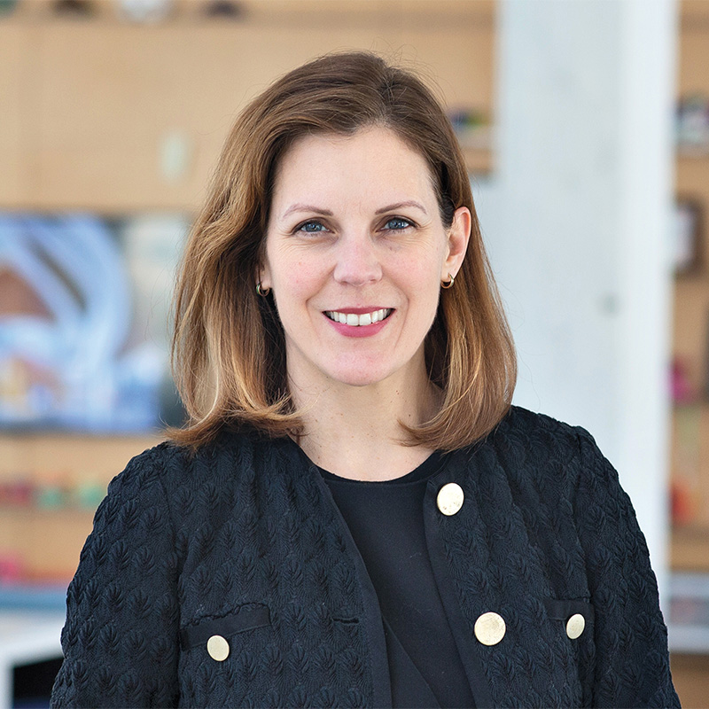 A woman with shoulder-length light brown hair, wearing a textured black jacket with gold buttons, stands indoors and smiles at the camera.