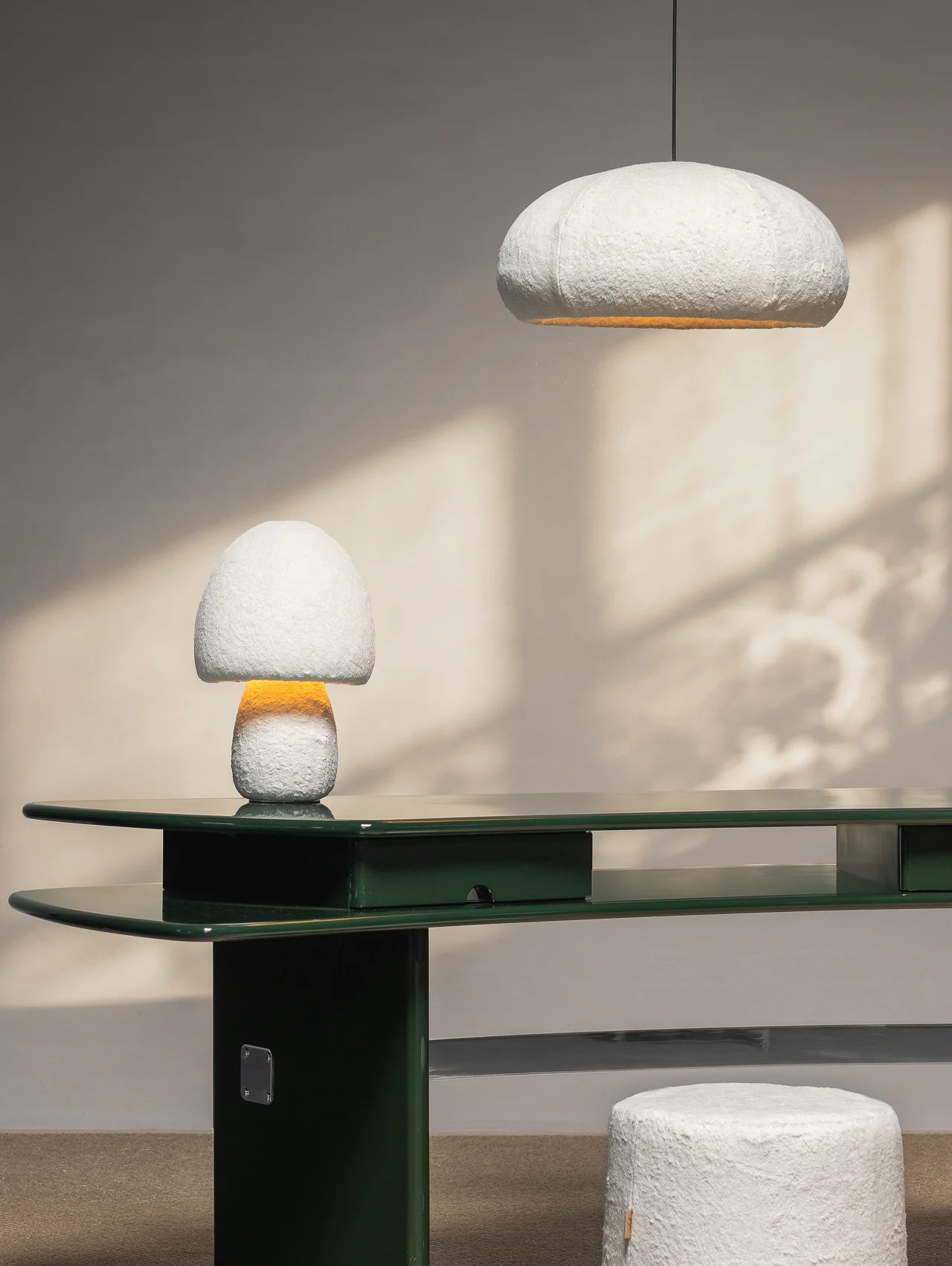 A modern green desk with a white mushroom-shaped lamp, matching ceiling light, and a round white stool in a minimalist room inspired by Milan Design Week 2026.