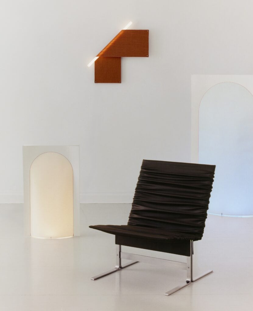 Minimalist room with a modern black chair, a white rectangular panel featuring an arch cutout, and an angular orange wall-mounted light—an inspired setting reminiscent of Milan Design Week 2026 style on a crisp white background.
