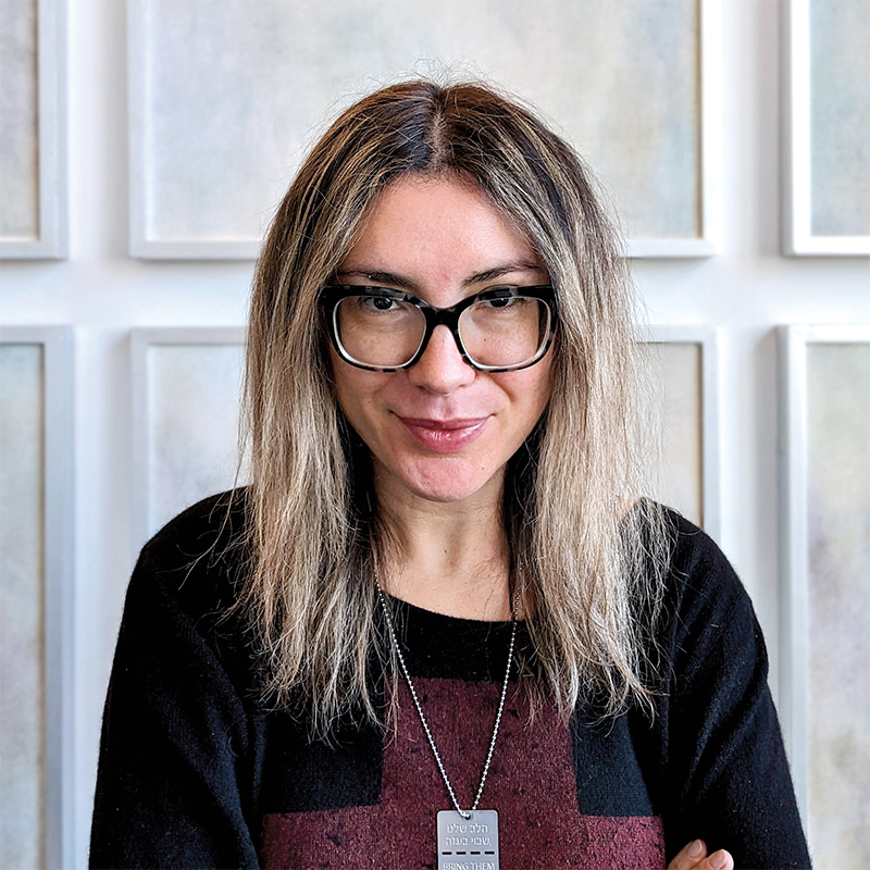 Person with long, light brown hair and black glasses, wearing a black and burgundy top and a rectangular pendant, standing in front of a light-colored wall with framed panels.