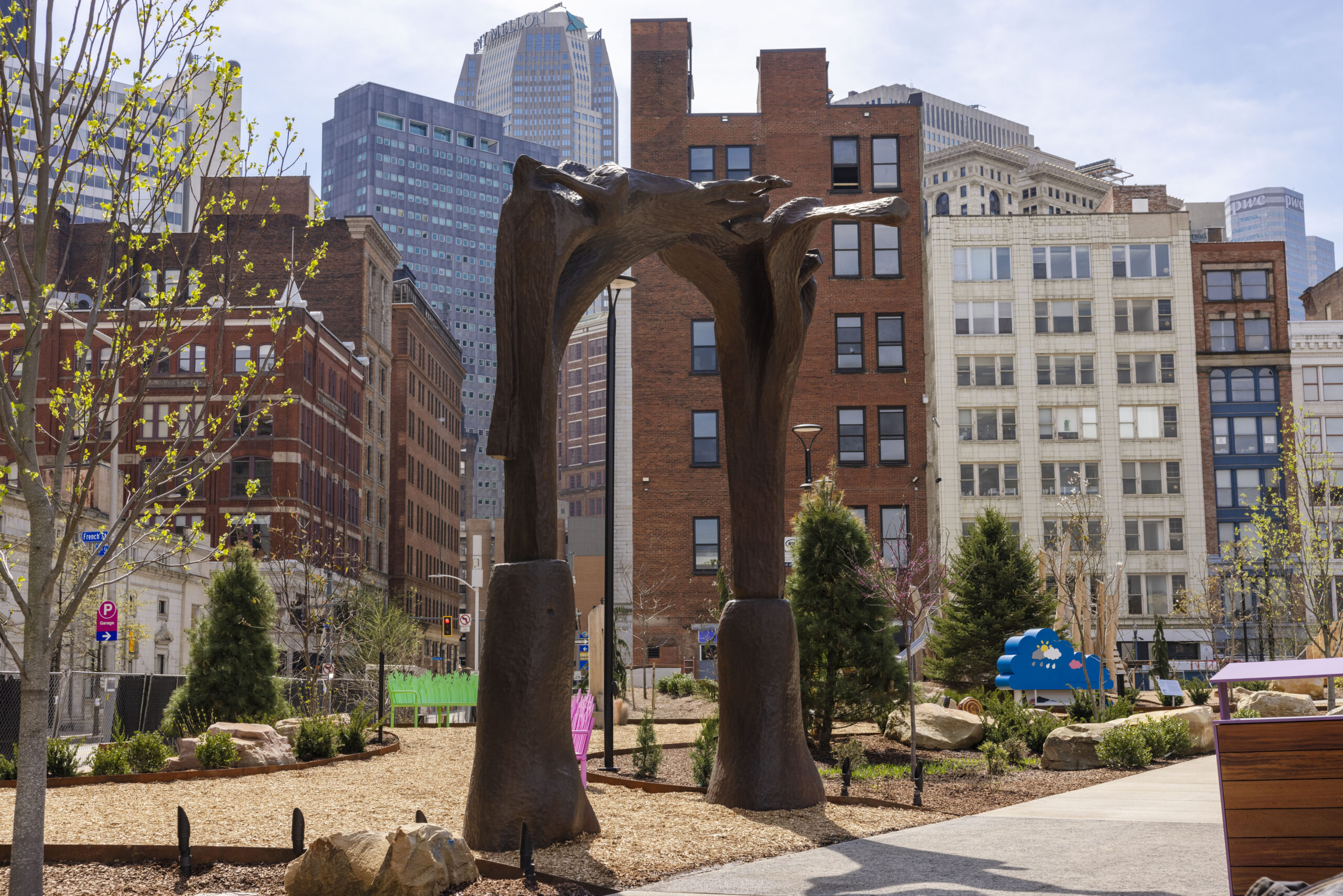 Large bronze sculpture with two arching forms stands in an urban park—landscaped by Field Operations—surrounded by trees, plants, rocks, and tall city buildings in the background.