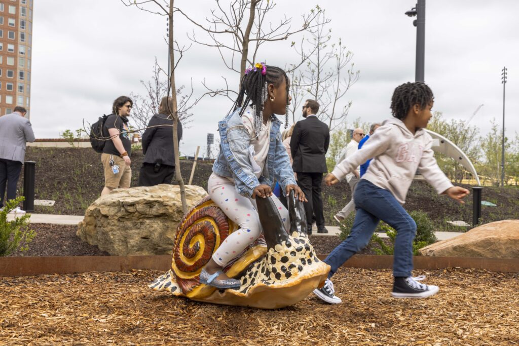 Two children play on a snail sculpture designed by Field Operations in an outdoor park, while several adults walk and converse in the background.
