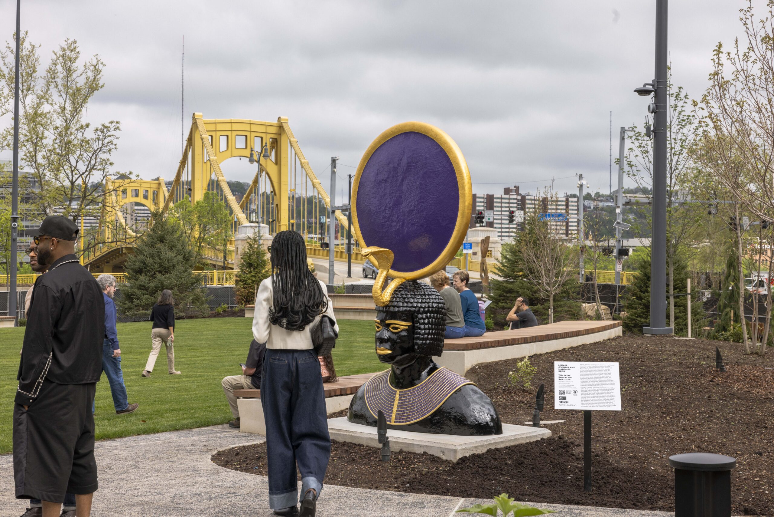 People walking near a large decorative sculpture created by Field Operations, featuring a head with a circular purple and gold element in a park, with a yellow bridge and city buildings in the background.
