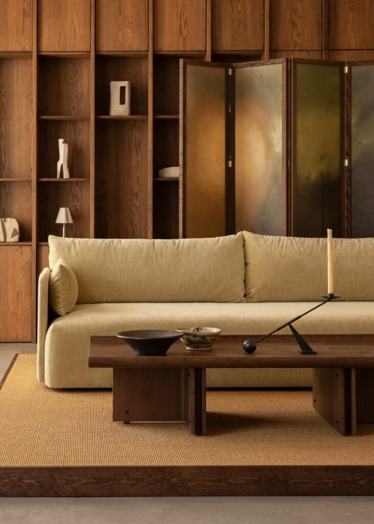 A beige sofa sits in front of a dark wood shelving unit with abstract decor; a wooden coffee table and neutral rug complete this modern living room, inspired by trends previewed at Milan Design Week 2026.