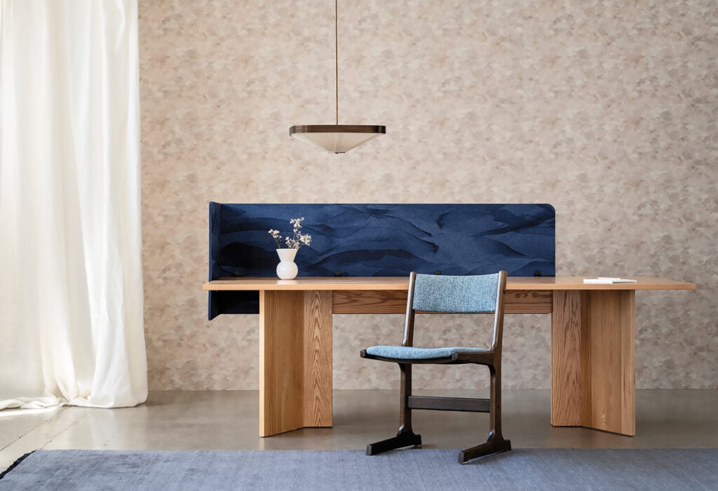 Minimalist office setup with a wooden desk, blue-cushioned chair, METROPOLISLikes blue fabric divider, white vase with flowers, pendant light, and textured beige wallpaper.