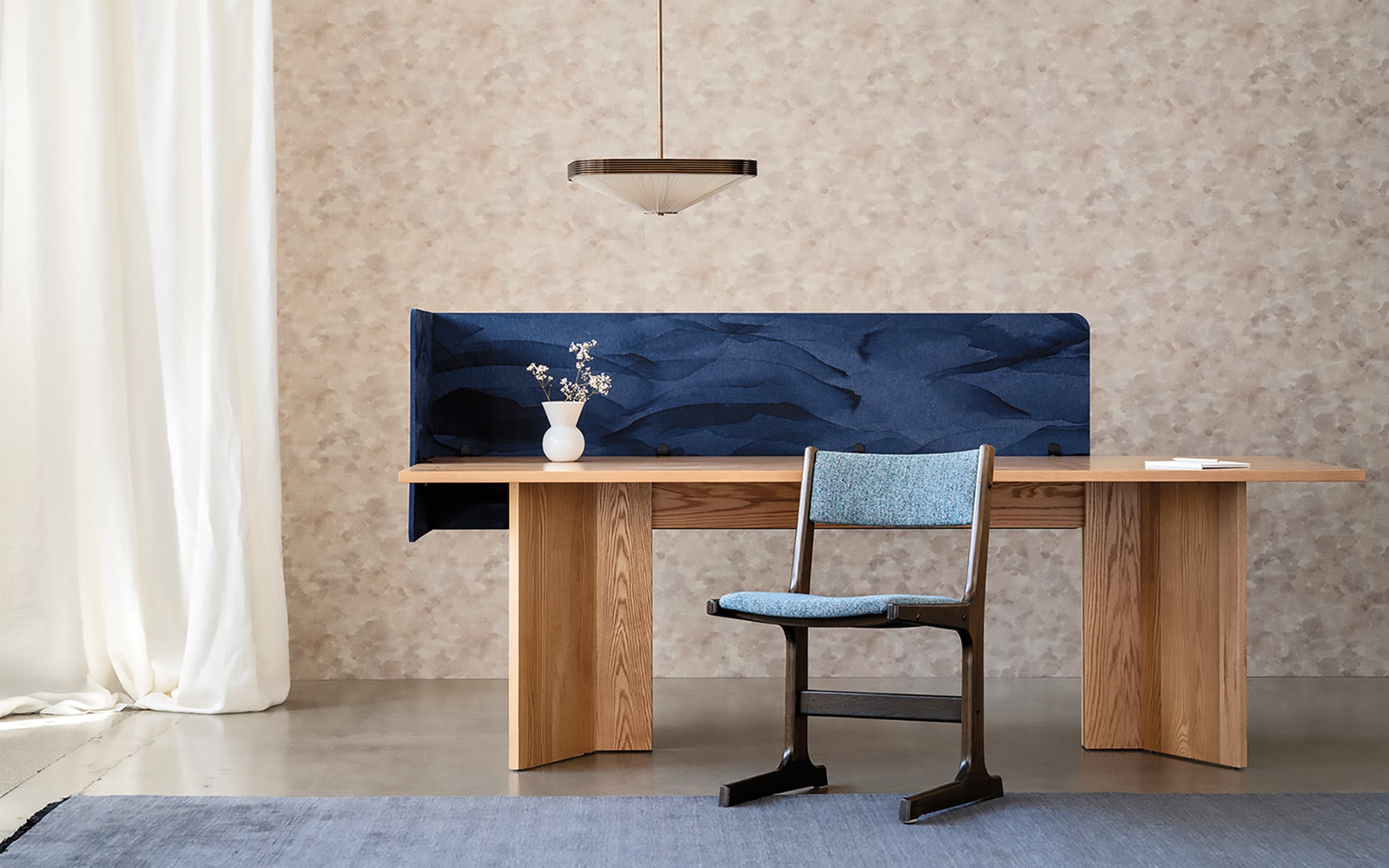Office products: A modern wooden desk with a blue fabric privacy panel, a blue-cushioned chair, a pendant light, and a white vase with flowers against a patterned wall and white curtain.