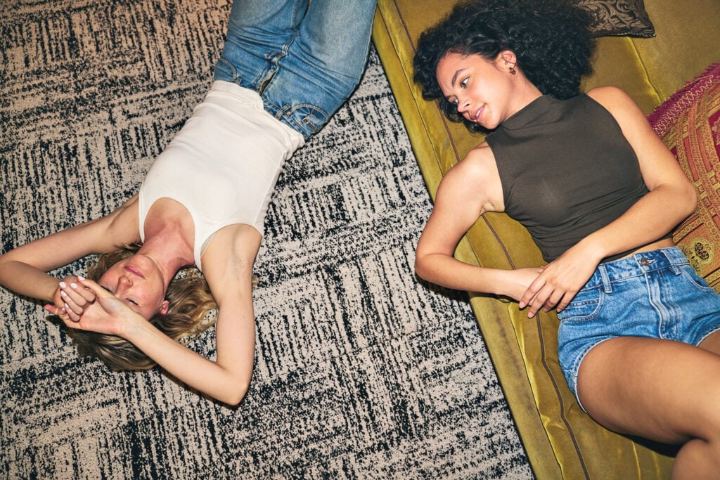 Two women are lying on a patterned carpet next to a yellow couch, facing each other and relaxing. METROPOLISLikes the cozy scene: one wears jeans and a white tank top, the other denim shorts and a sleeveless dark top.