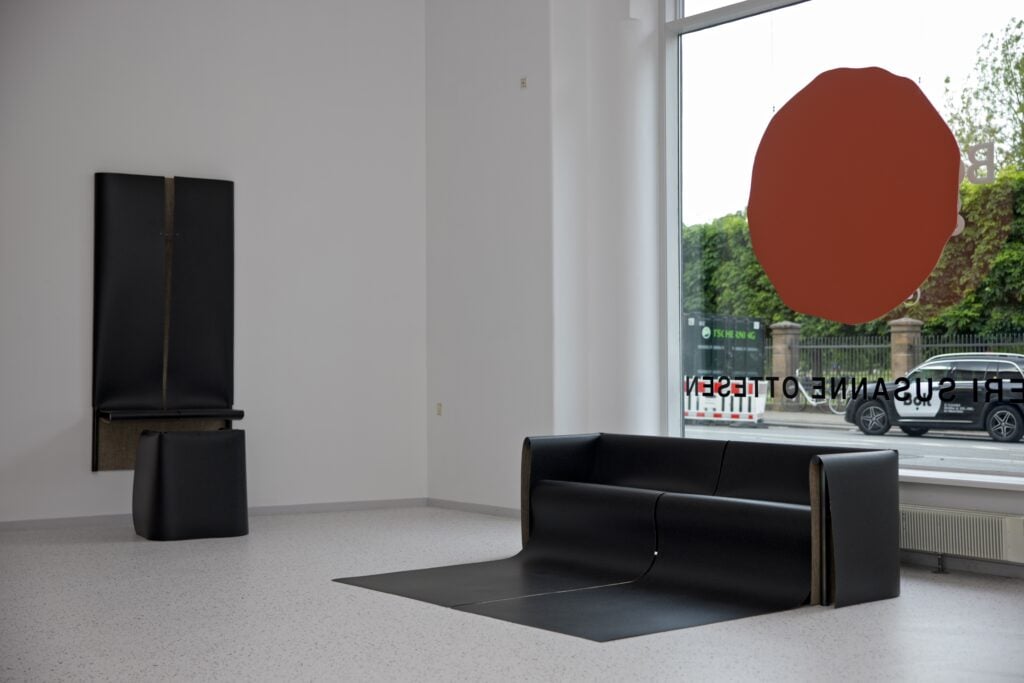 Minimalist interior with a black couch and matching chair, large window featuring a circular orange decal, and greenery visible outside—an aesthetic inspired by trends previewed at Milan Design Week 2026.