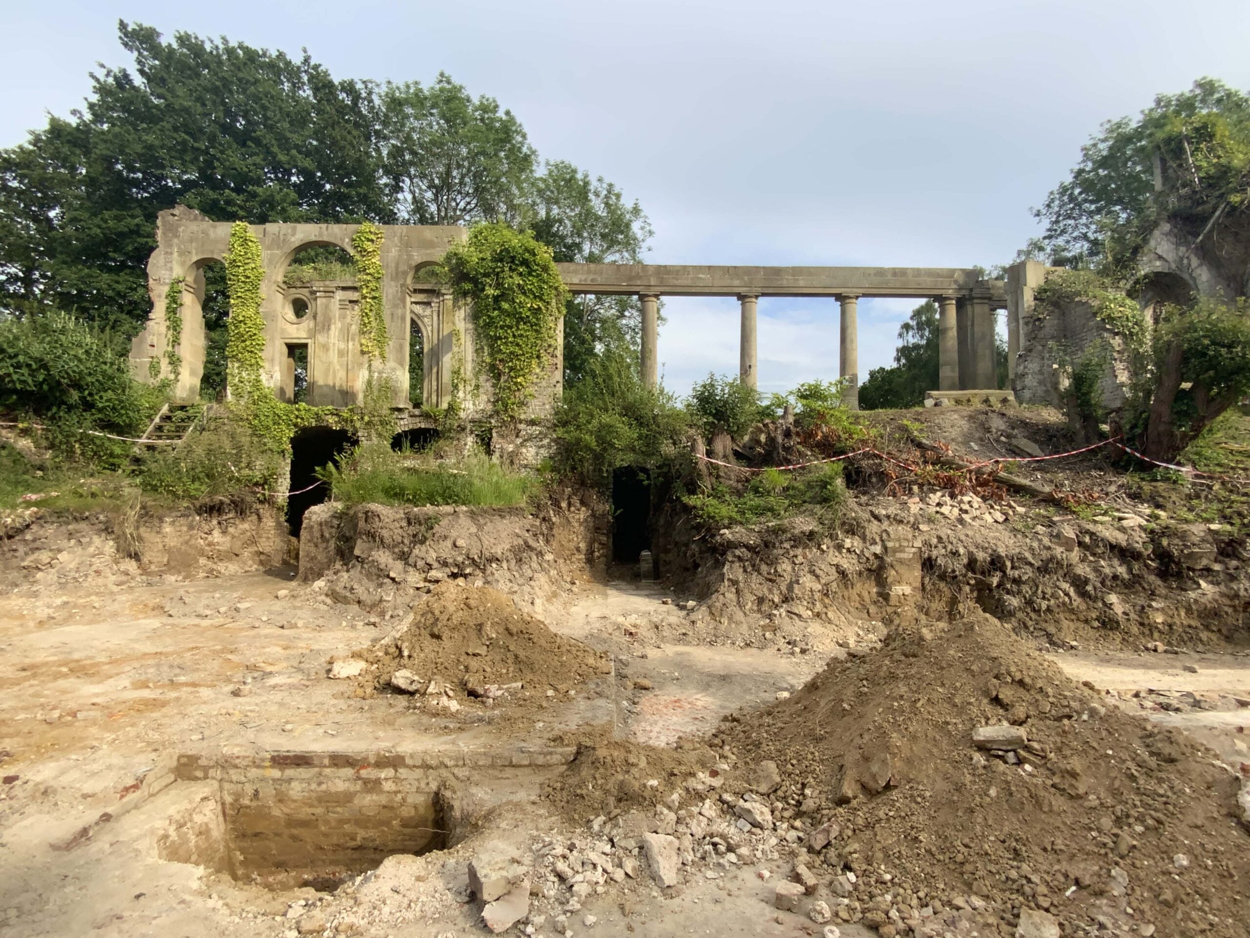 Ruins of an old stone building with arches and columns, partly covered in ivy, surrounded by excavated dirt and debris under a cloudy sky. Stone fragments lie scattered among the remains.
