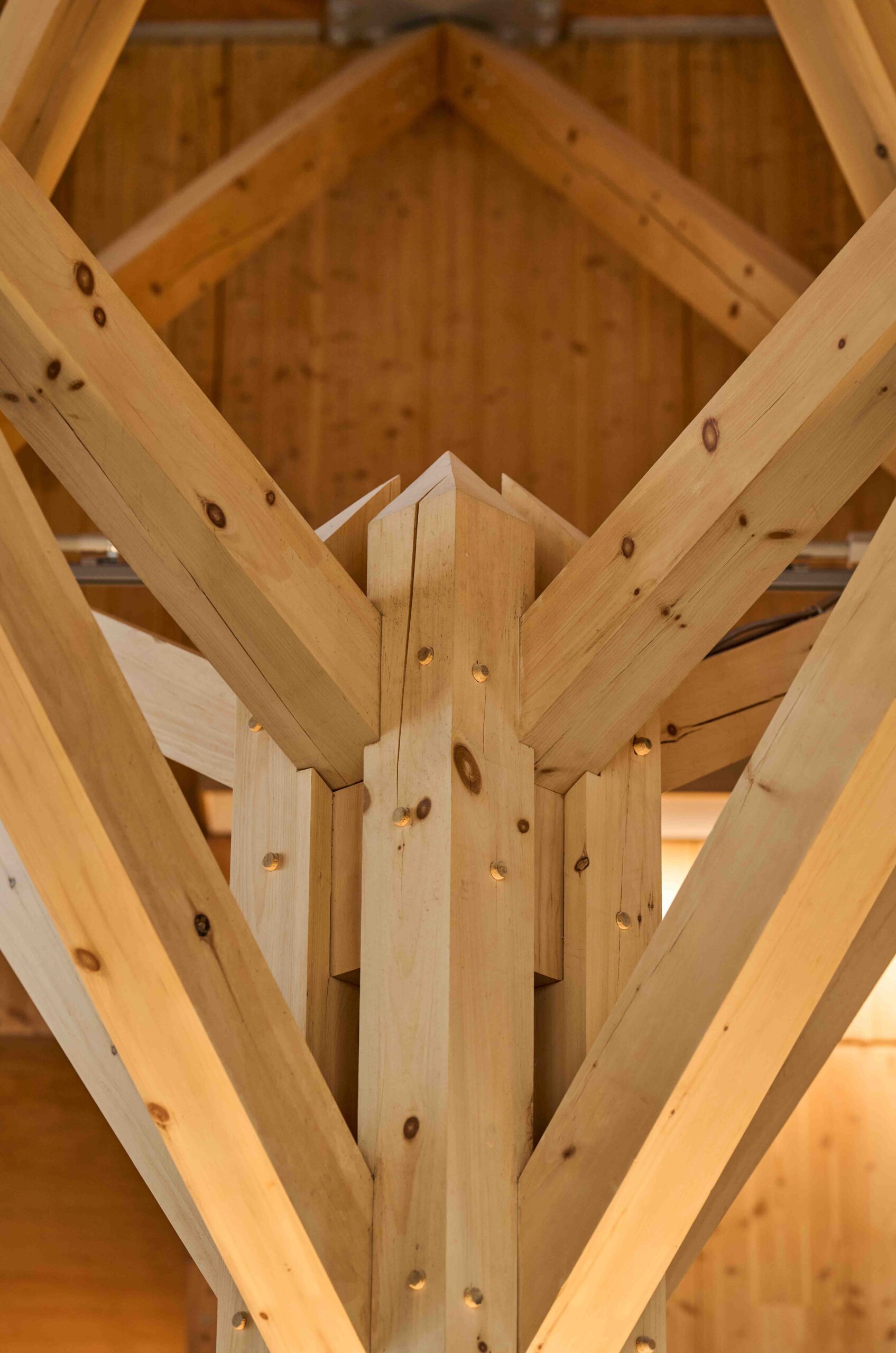 Close-up of an intersecting timber joint structure with light wooden beams, showing detailed craftsmanship and exposed bolts inside a wooden building.