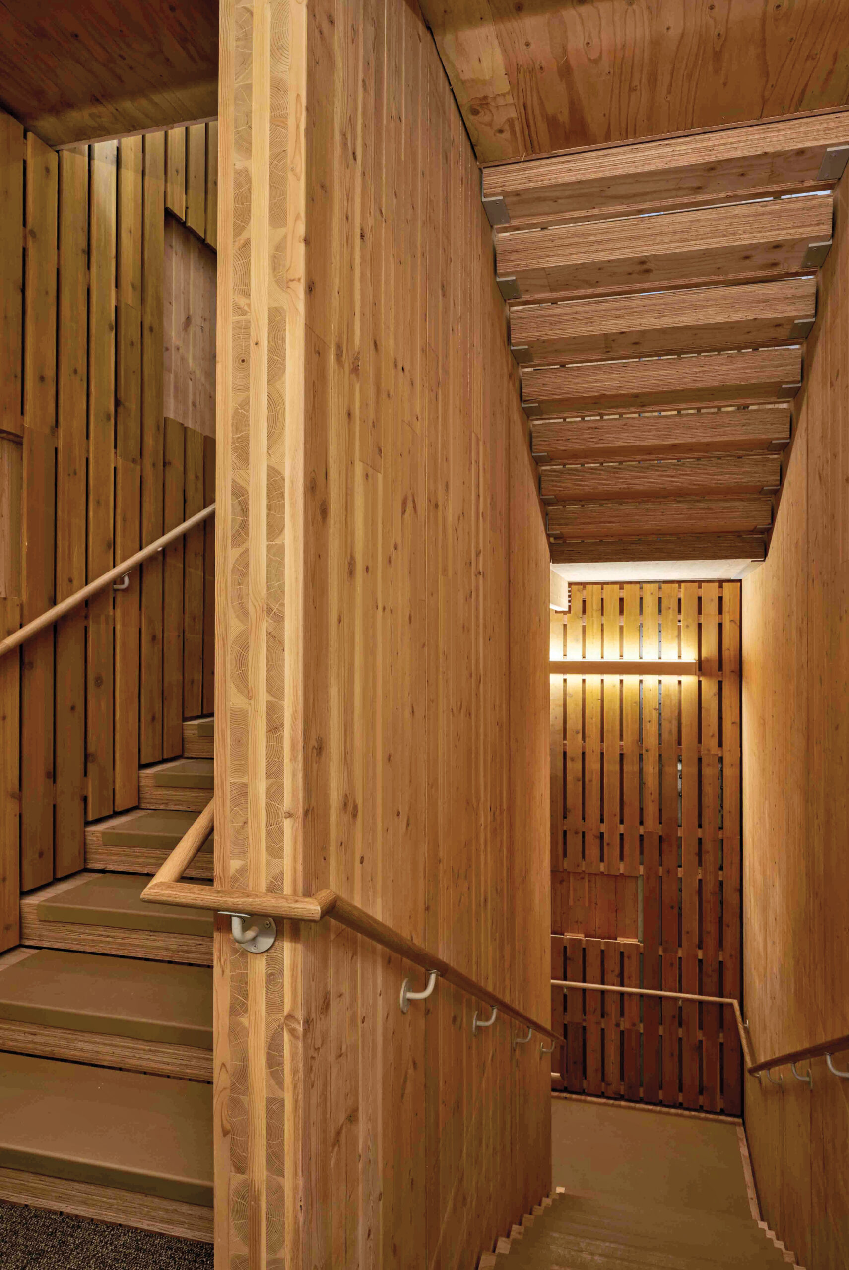 Indoor staircase and walls made entirely of light wood with vertical slats, handrails on both sides, and lighting mounted on the wall.