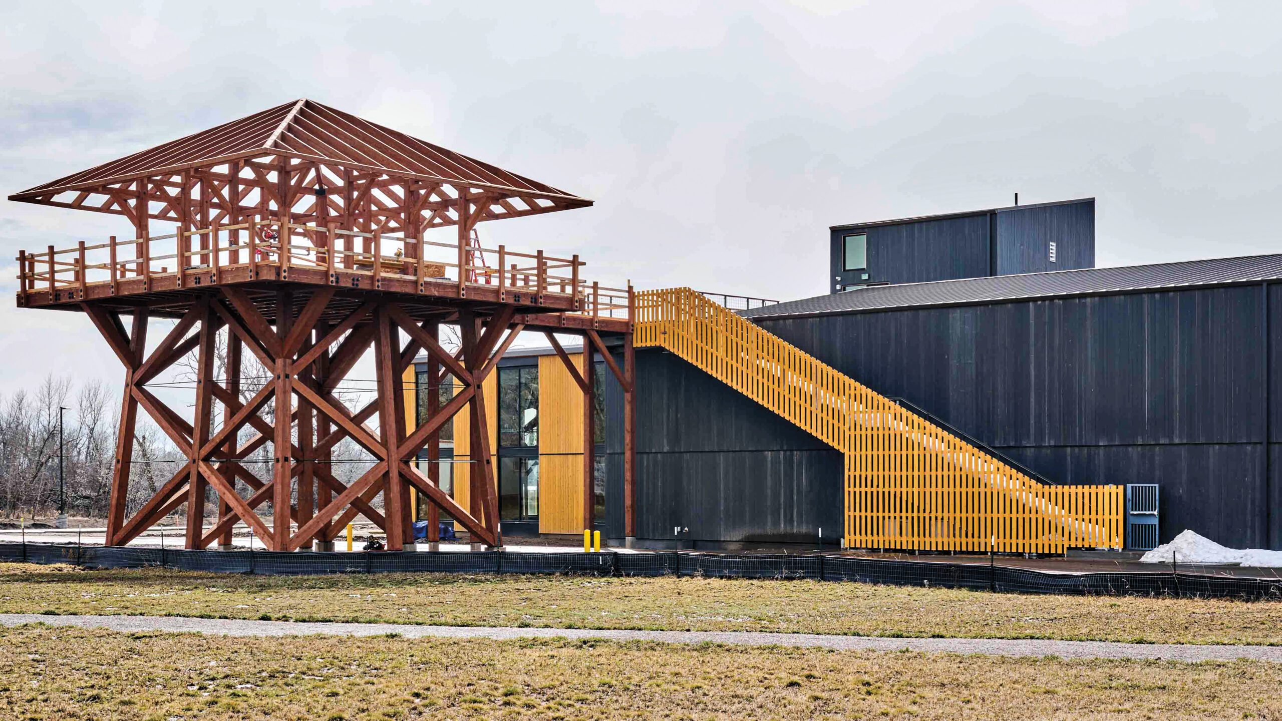 Red timber structure and bright yellow staircase made of wood slats and black building in a snowy, grass landscape.
