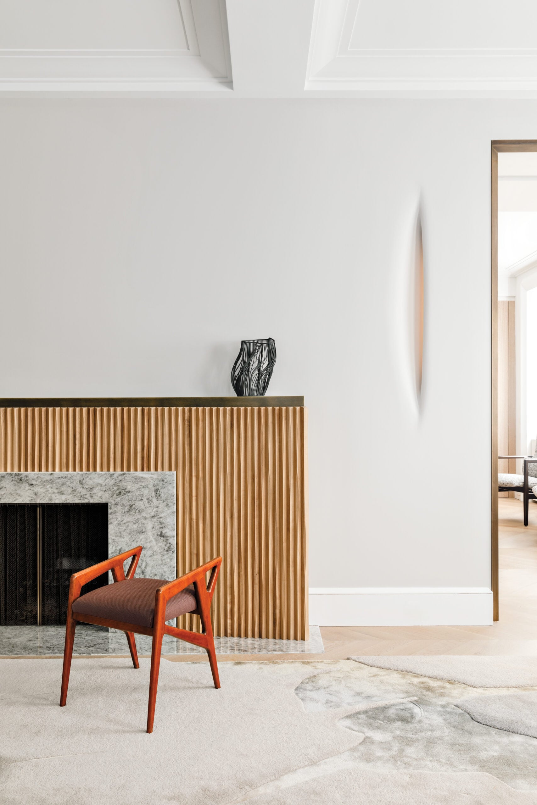 A modern room with a marble and wood fireplace, a sculptural black vase on the mantel, a red chair, and a light-colored abstract rug on the floor.