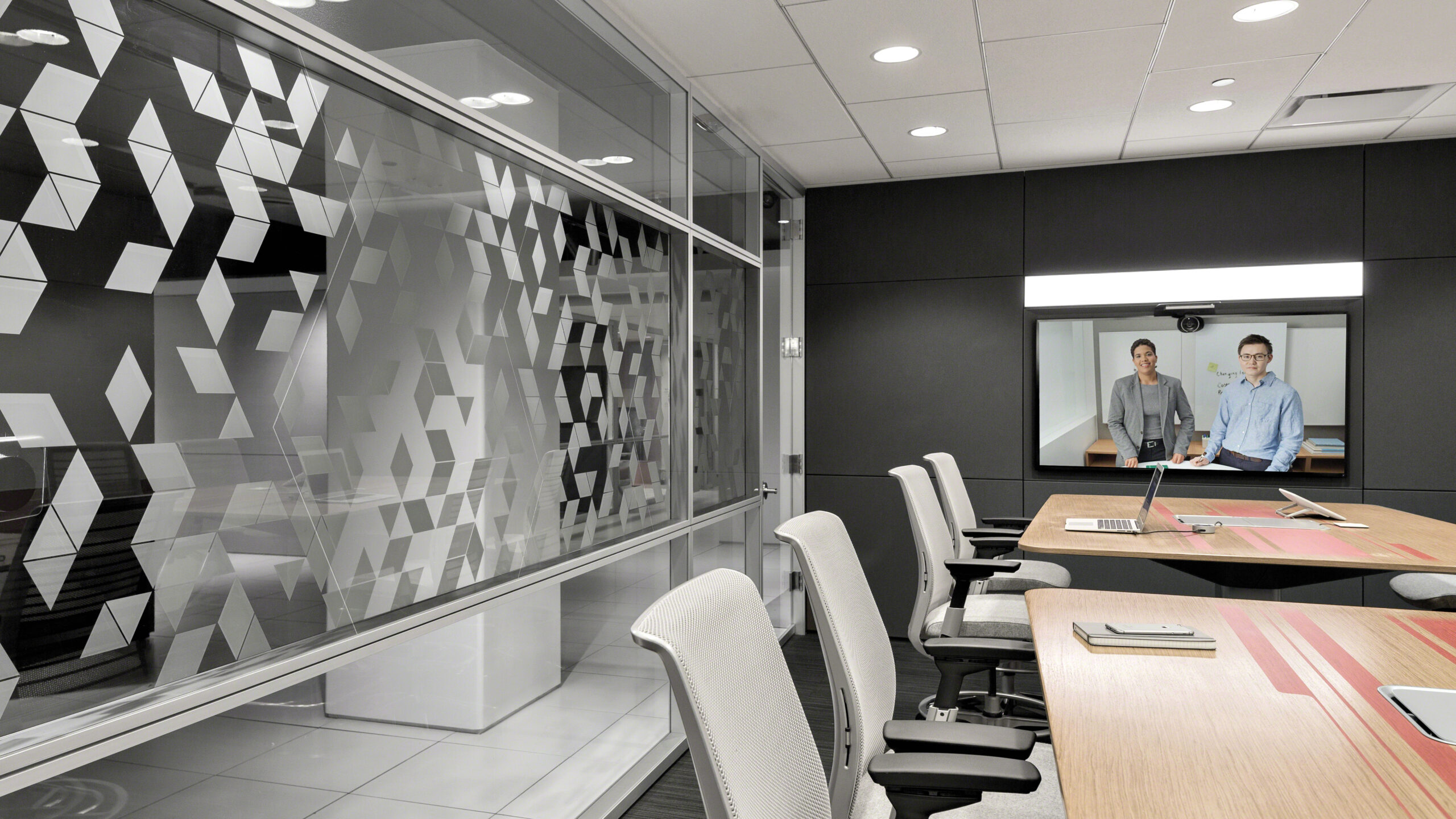 Modern conference room with a geometric-patterned glass wall, empty chairs, laptops on the table, and two people on a video screen at the far end of the room.