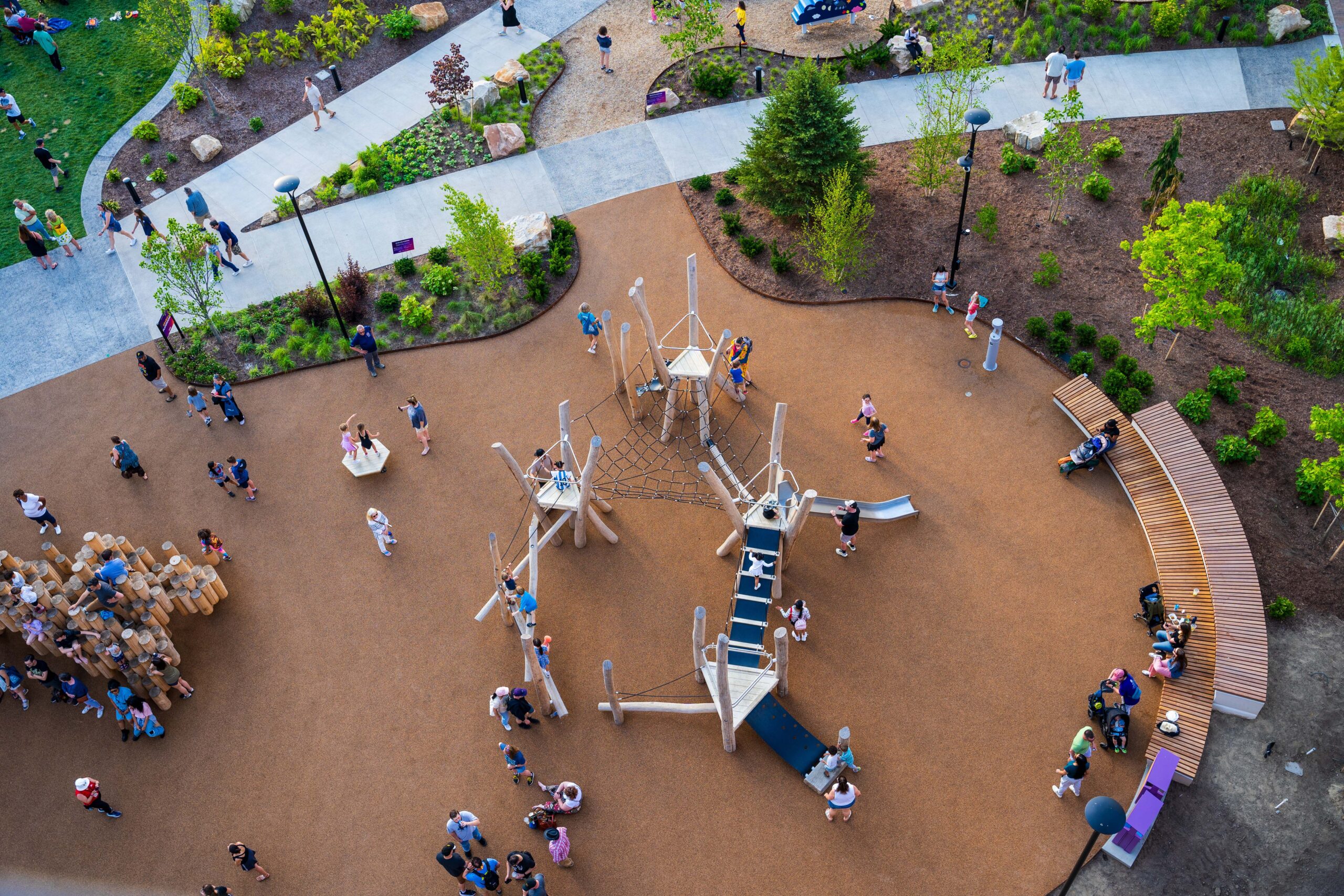Aerial view of a playground with children and adults scattered around play structures, paths, benches, and landscaped areas on a sunny day.