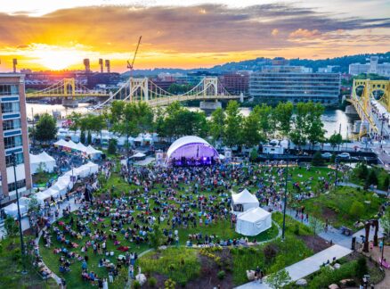 Outdoor concert with a large crowd seated on grass in a park, tents set up, and bridges spanning a river at sunset in an urban setting.