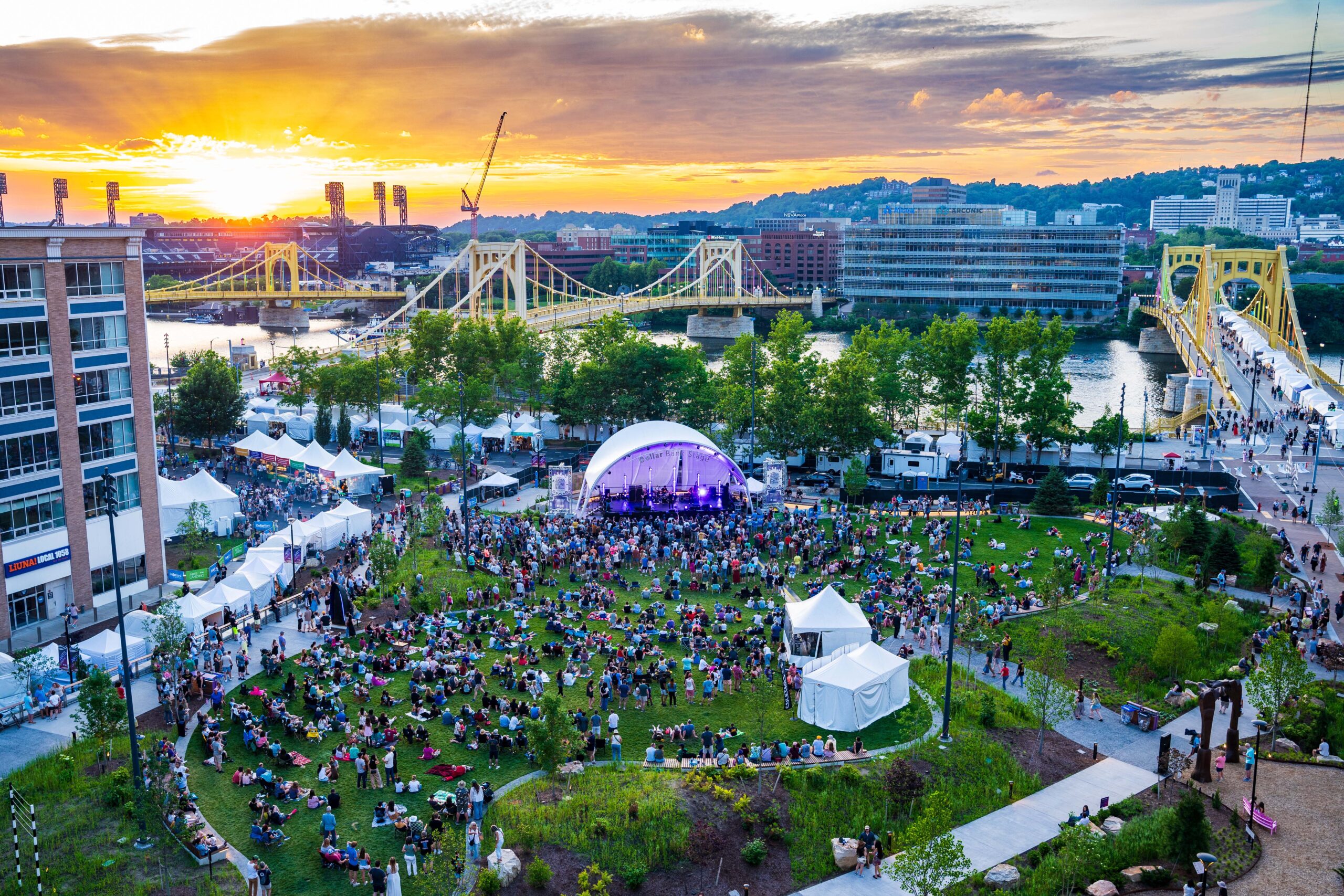 Outdoor concert with a large crowd seated on grass in a park, tents set up, and bridges spanning a river at sunset in an urban setting.