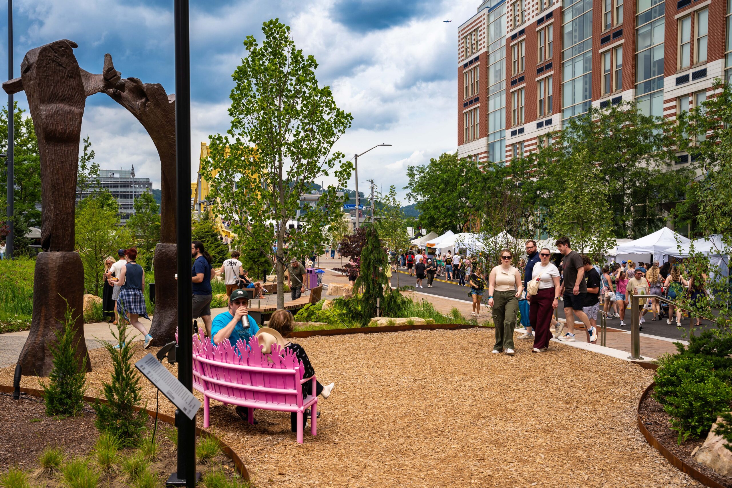 People walk through a busy outdoor festival with vendor tents, while others sit on a pink bench in a landscaped park area. Tall buildings and sculptures are visible in the background.