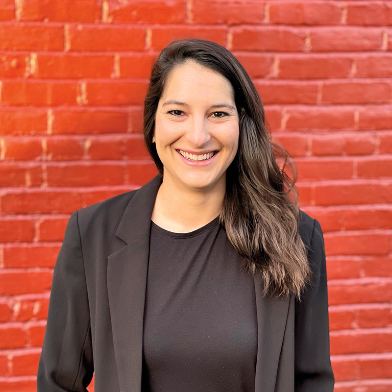 A woman with long brown hair wearing a black blazer and top stands smiling in front of a red brick wall.