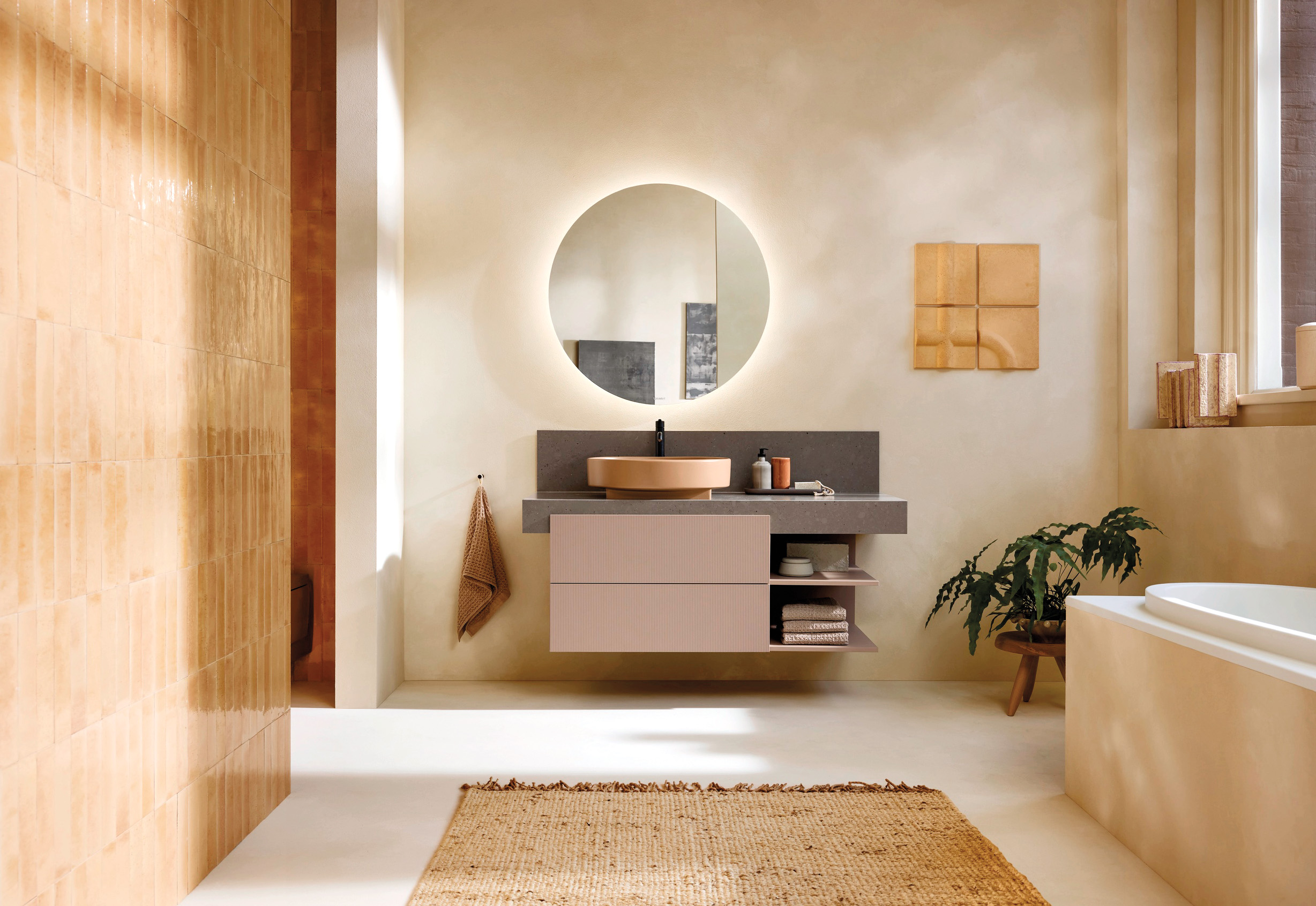 Modern bathroom with a round backlit mirror, floating vanity, vessel sink, bathtub, potted plant, wall tiles, and a woven rug on a light floor. Natural light streams in from the window.