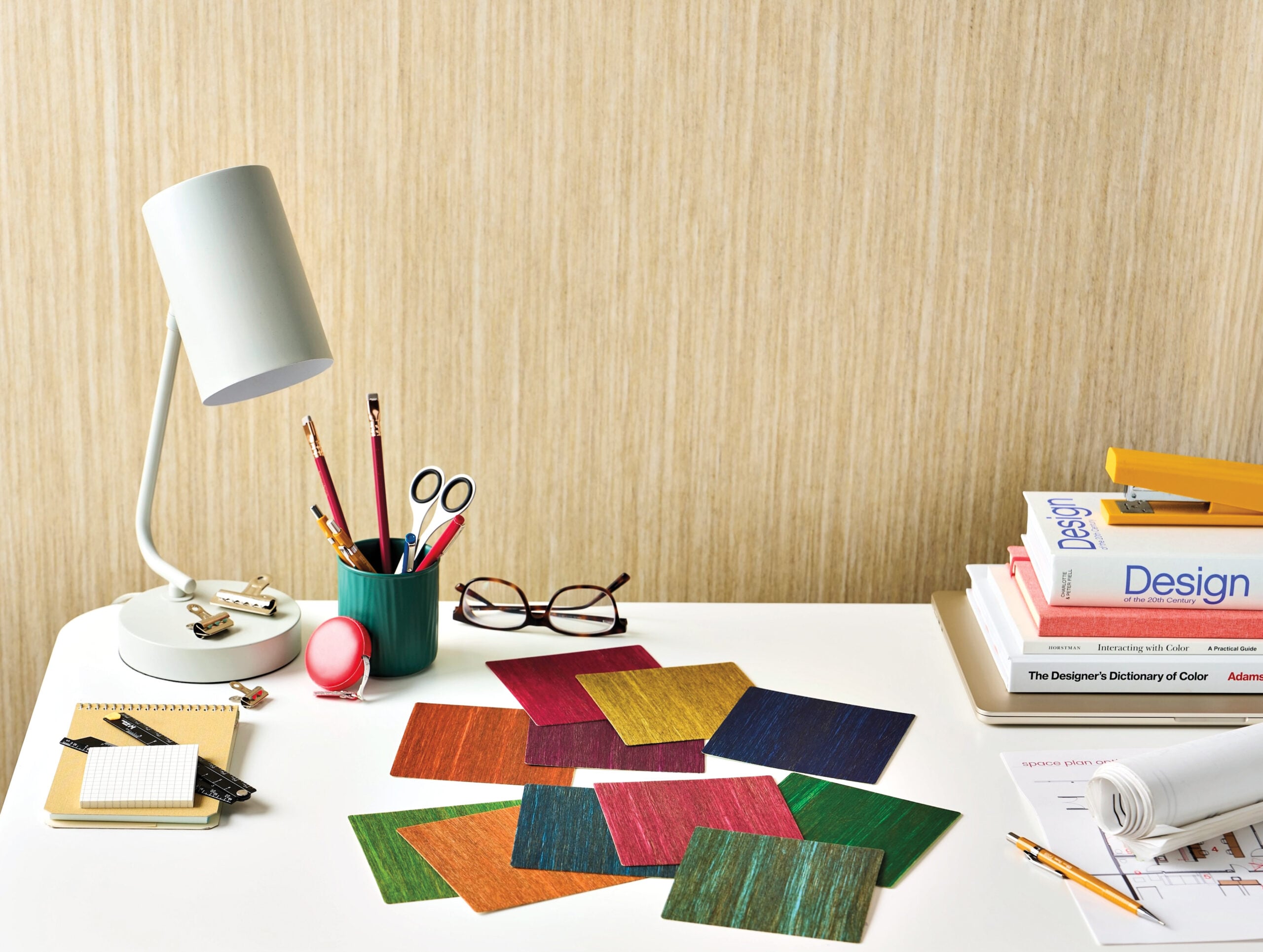 A white desk with colored pencil holders, scattered colored swatches, a lamp, books on design, eyeglasses, and office supplies against a beige wall.