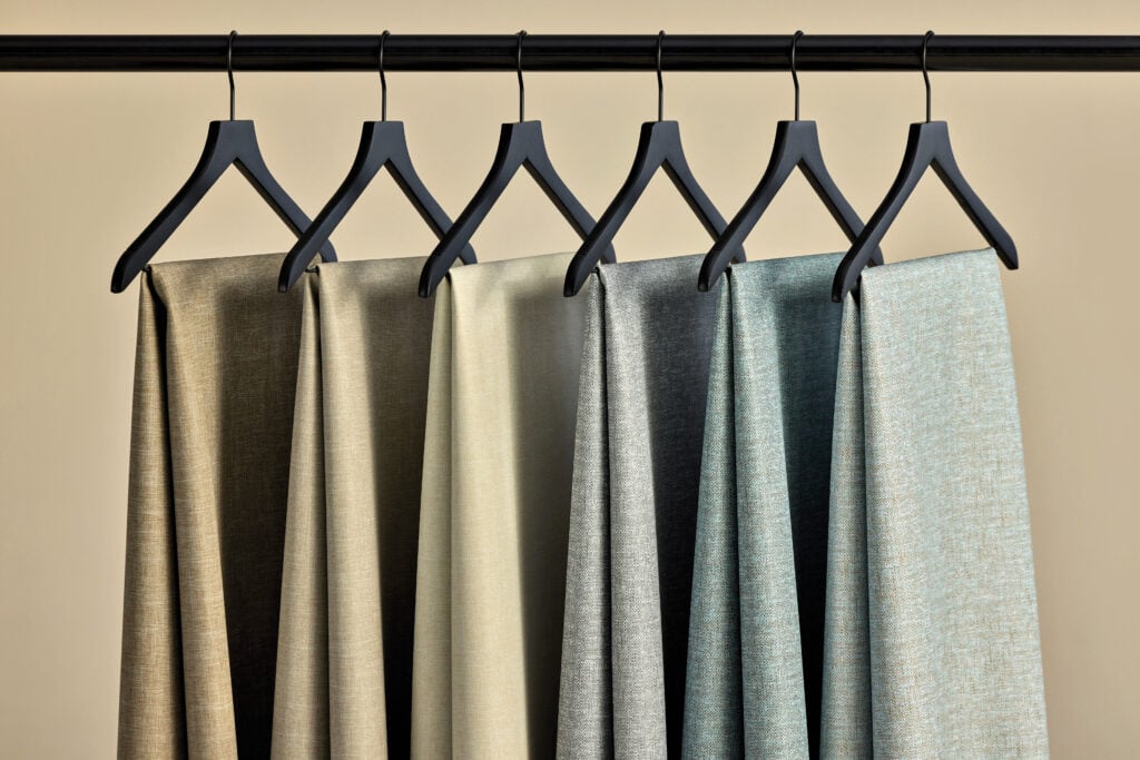 Six fabric panels in neutral and pastel shades hang on black hangers from a black rod against a beige background, showcasing timeless style that METROPOLISLikes.