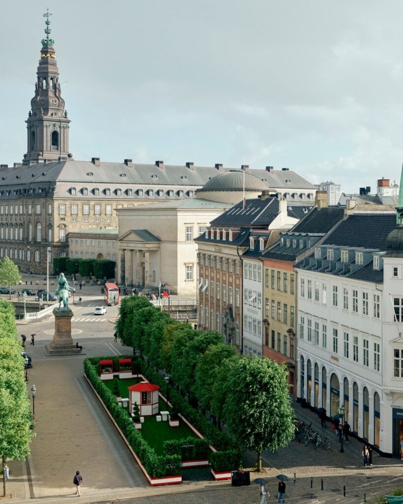 A cityscape with historic buildings, a statue in a plaza, a row of trees, and a green park area with red structures in the foreground under a cloudy sky, capturing the creative spirit of Milan Design Week 2026.