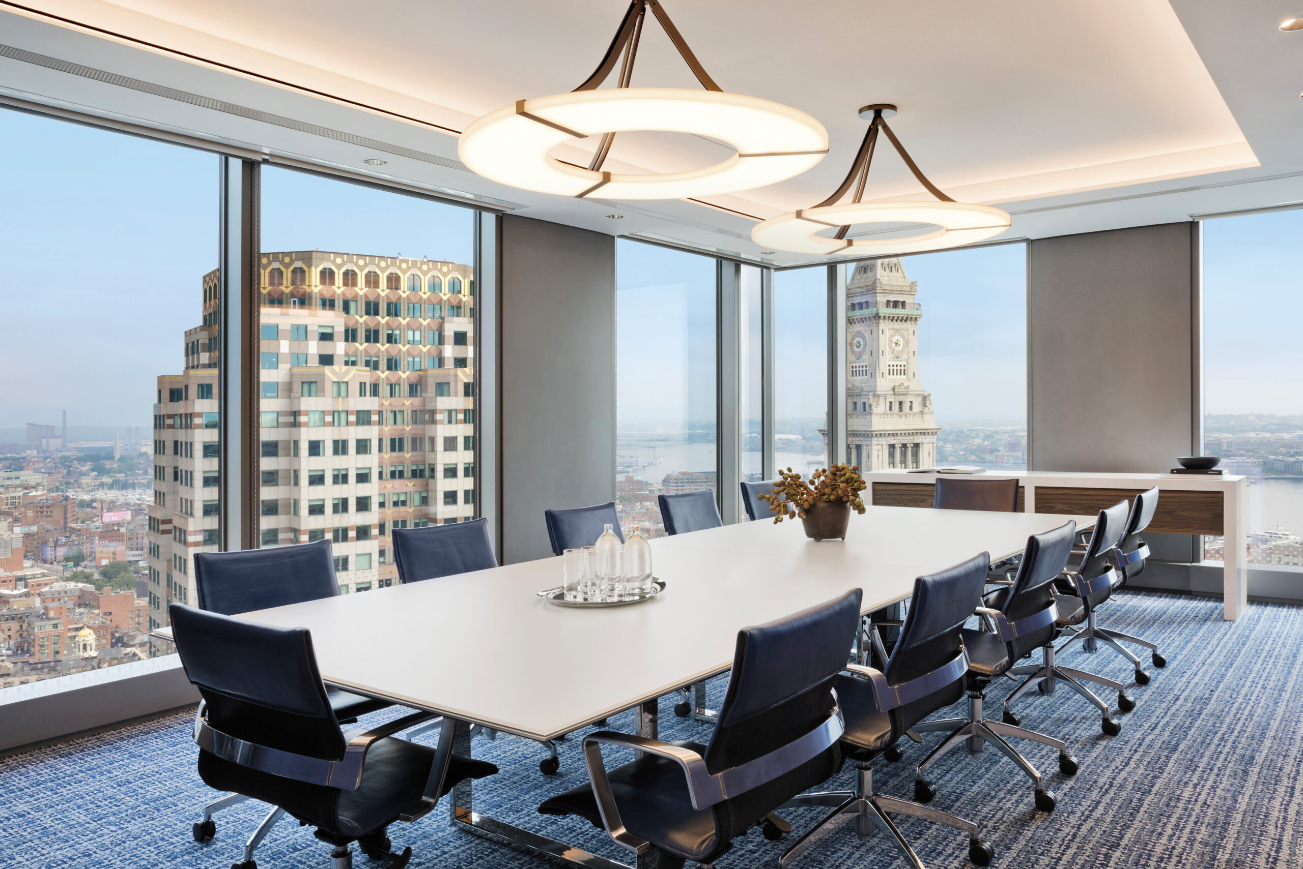 Modern conference room with a long white table, blue chairs, water glasses, large windows, and a cityscape view outside.