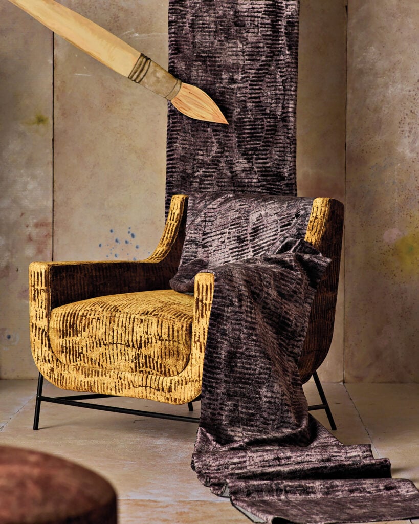 A textured yellow armchair is draped with dark brown fabric, with an oversized paintbrush suspended above in a neutral-toned room—a scene sure to capture METROPOLISLikes for its striking, modern aesthetic.