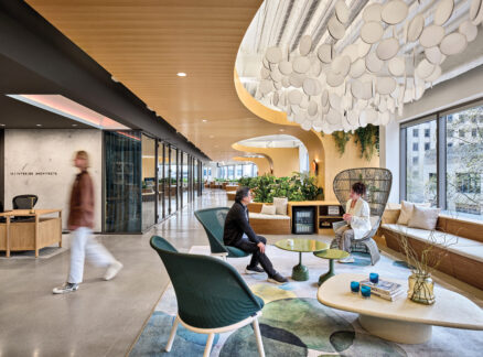 Modern Seattle office lobby with contemporary furniture, large round light fixture, and three people interacting; large windows provide natural light and greenery is visible inside and outside.