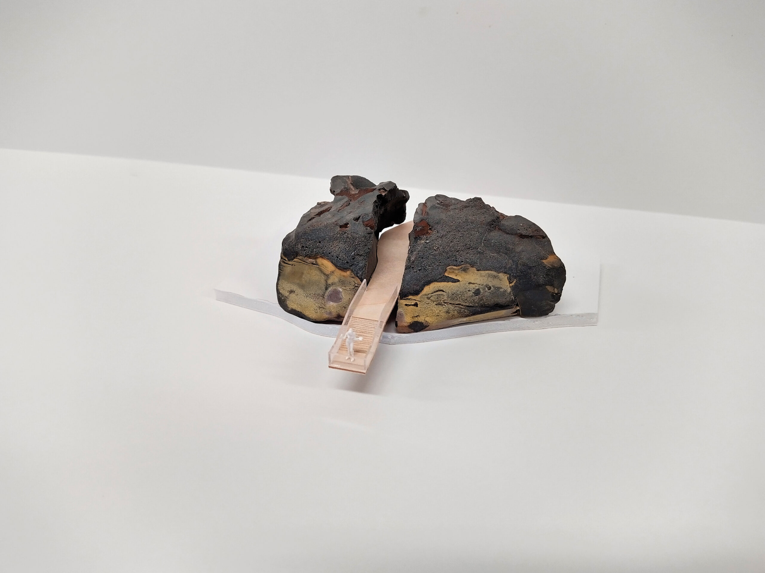 Two pieces of a dark rock are positioned on either side of a small beige object resembling a model bridge, all placed on a white surface.