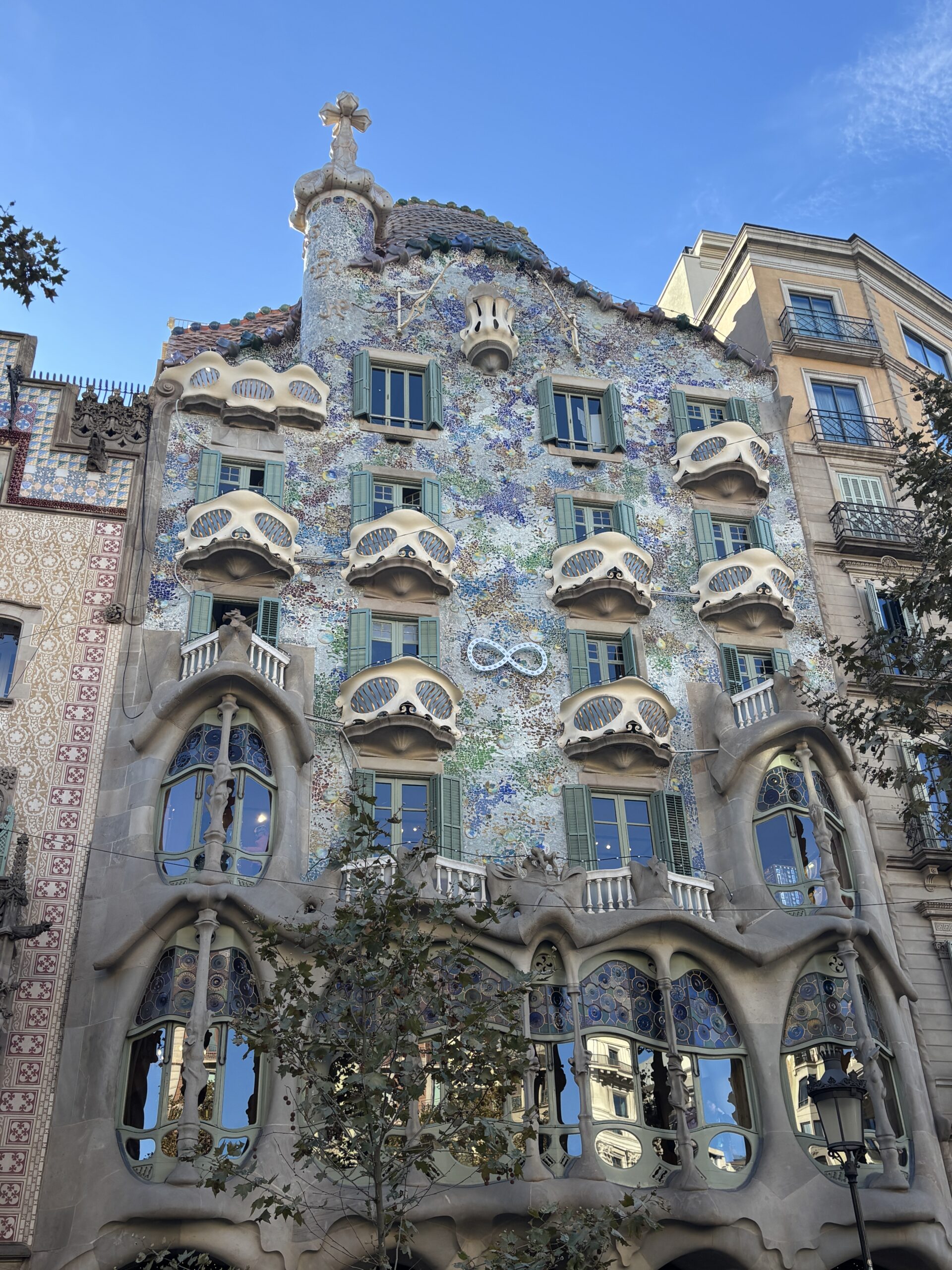 A view of the ornate façade of Casa Batlló in Barcelona, featuring colorful mosaics, wavy balconies, and irregularly shaped windows under a clear blue sky.